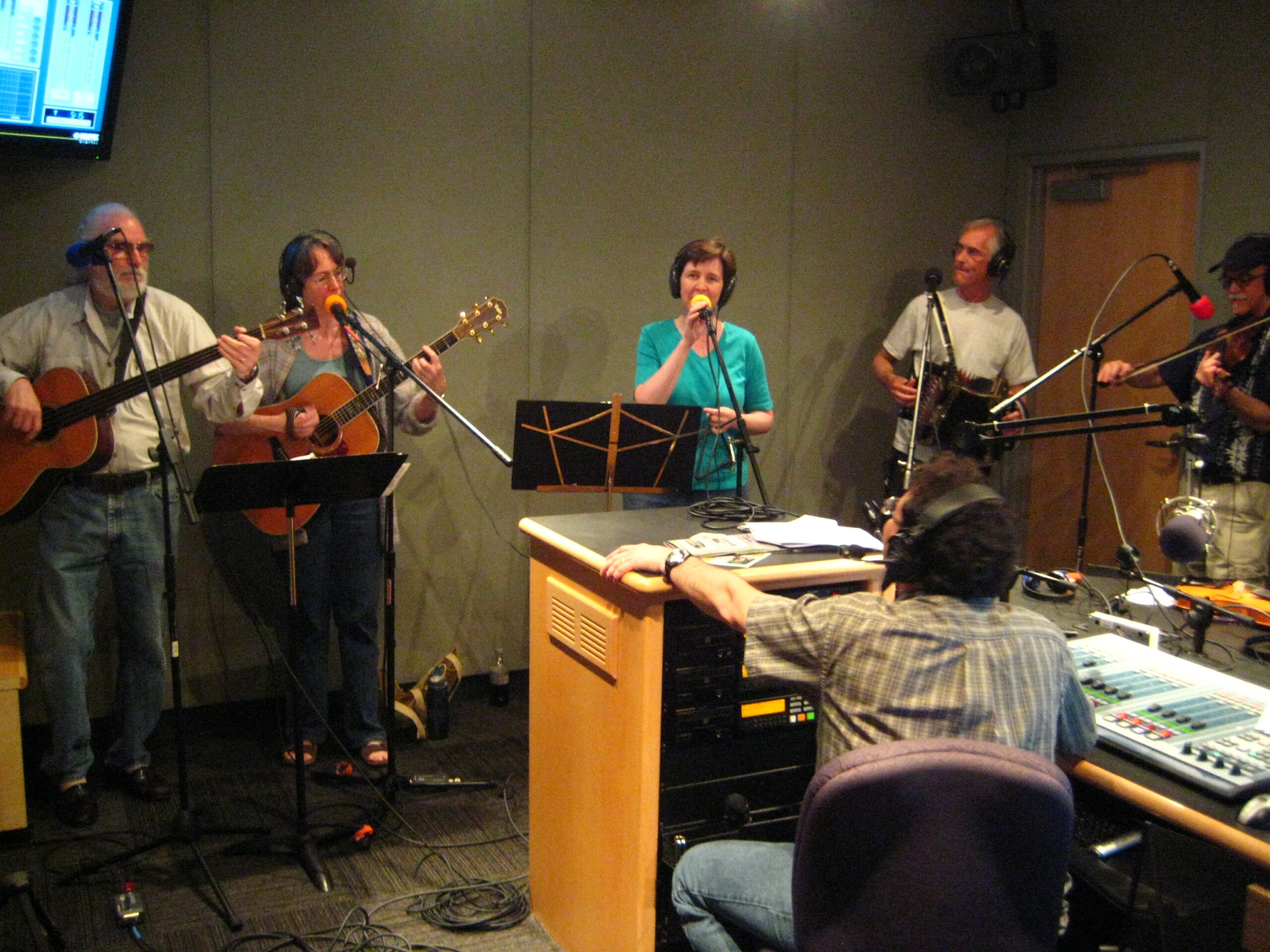 Two adults playing acoustic guitars, an adult singing into a microphone, an adult playing an accordion, and an adult playing a violin as an adult wearing headphones listens in a radio studio
