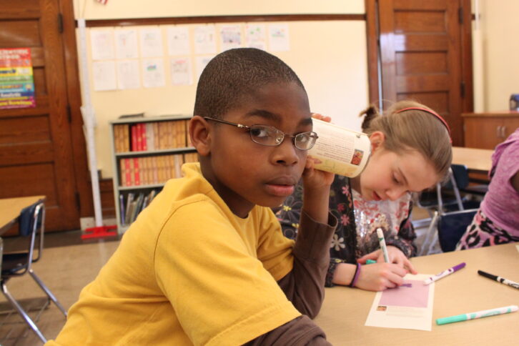A youth holding a can up to their ear as another youth draws on a piece of paper in a classroom