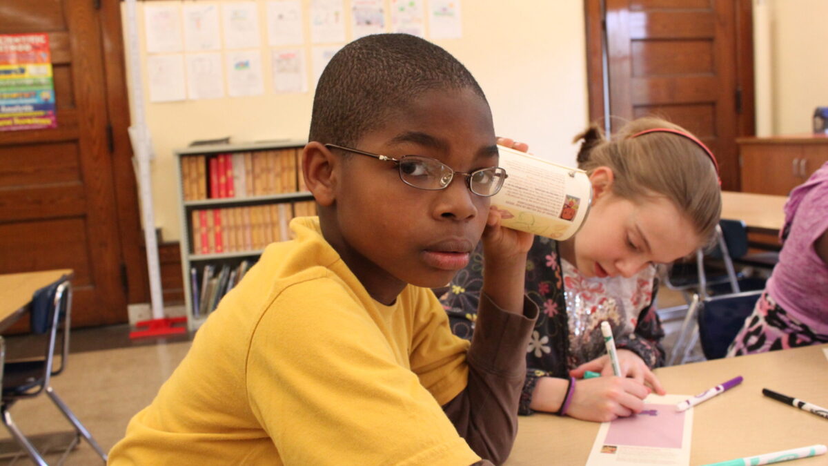 A youth holding a can up to their ear as another youth draws on a piece of paper in a classroom