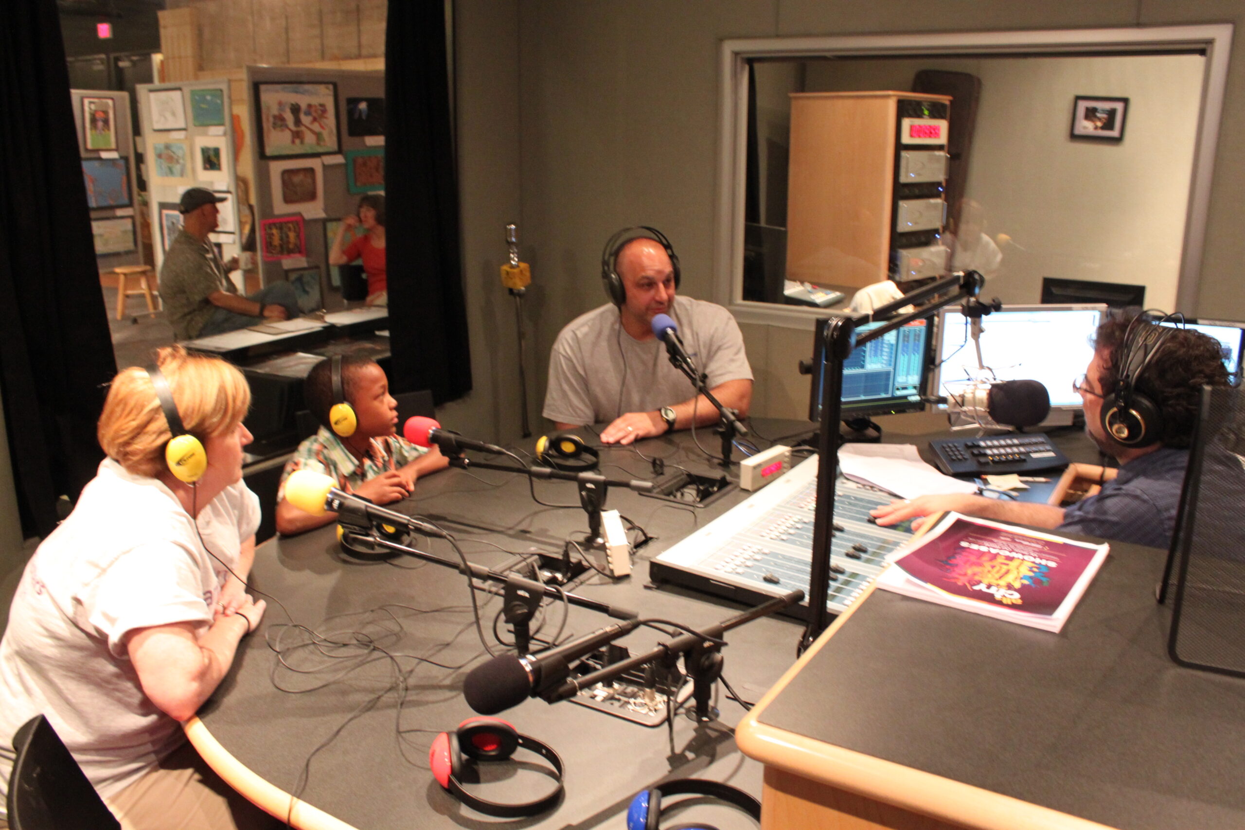 Two adults and a youth wearing headphones speaking into microphones being interviewed by an adult wearing headphones sitting behind a microphone and an audio mixer in a radio studio