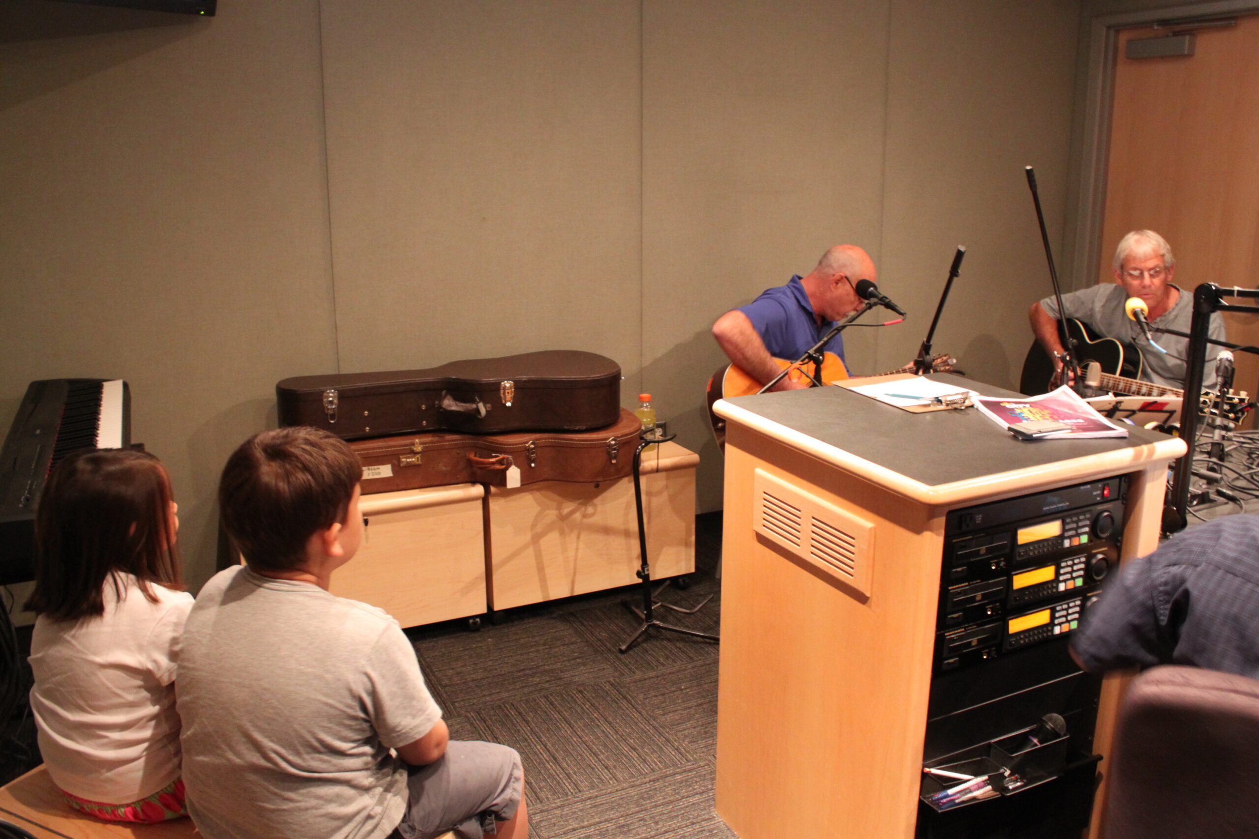Two adults playing acoustic guitars and two individuals watching in a radio studio