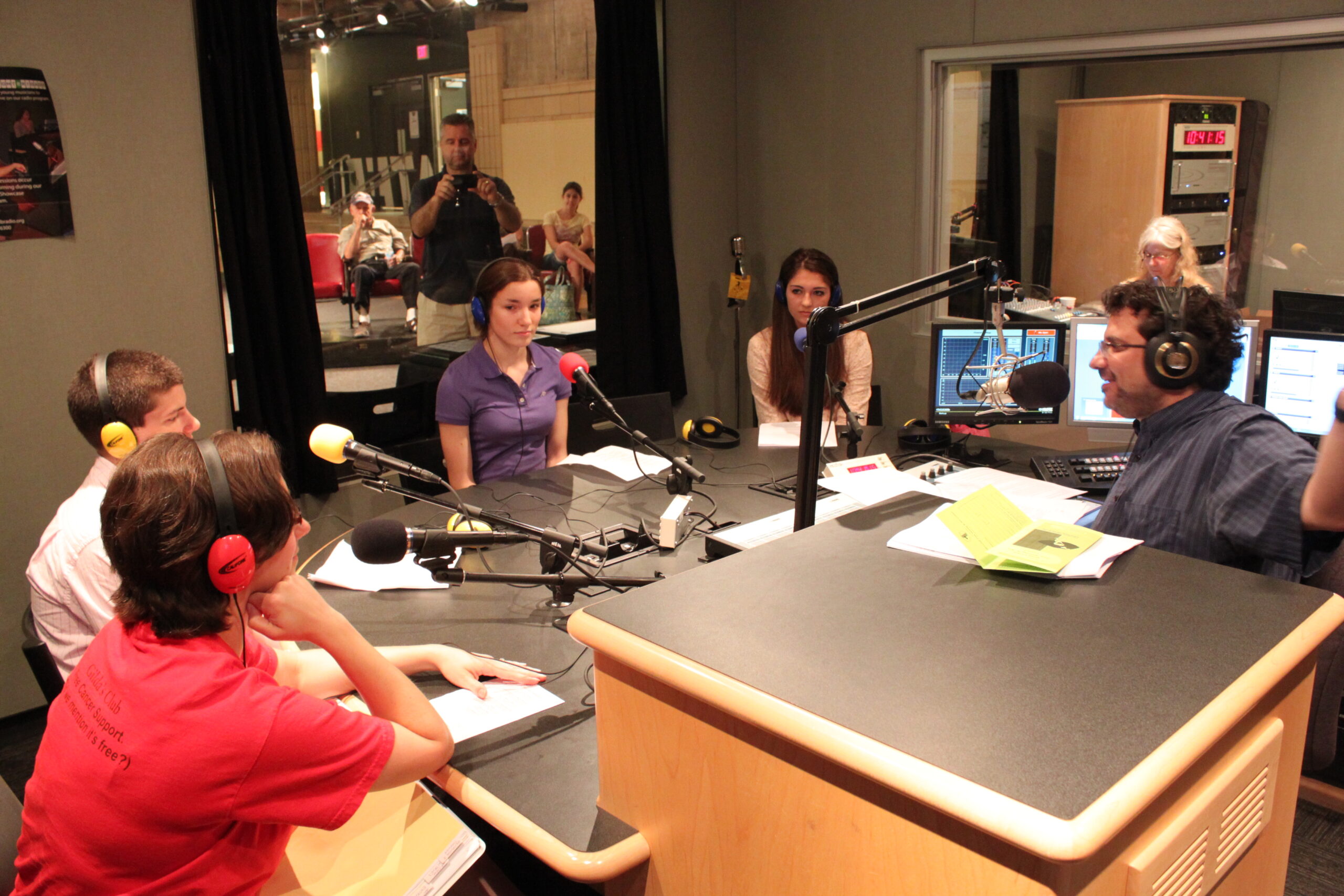Four teens wearing headphones speaking into microphones being interviewed by an adult wearing headphones sitting behind a microphone in a radio studio