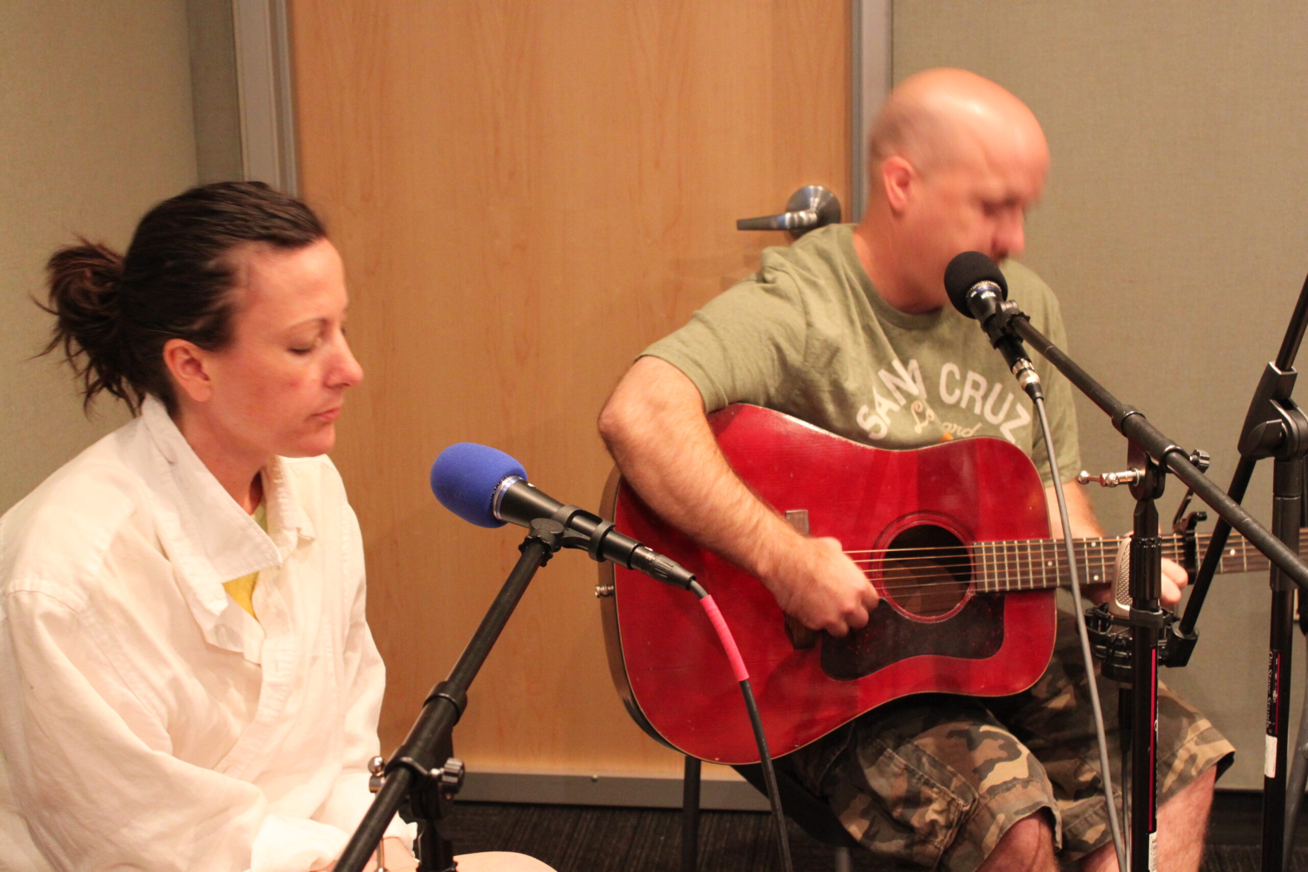 An adult sitting behind a microphone and an adult playing an acoustic guitar and singing into a microphone in a radio studio