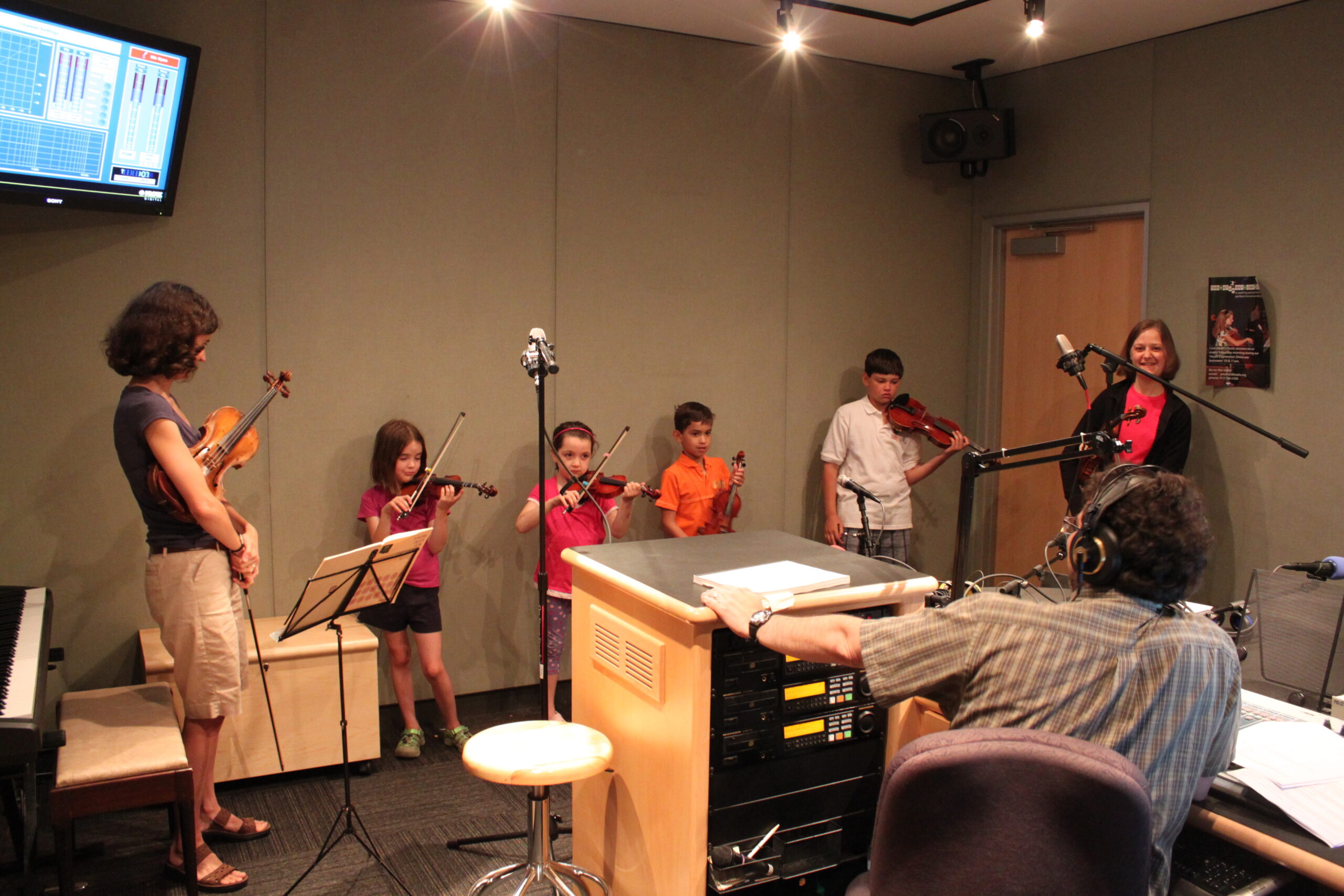 Five youth and an adult holding violins and an adult wearing headphones watching in a radio studio