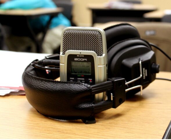 A pair of headphones around a portable recorder sitting on a wooden desk in a classroom