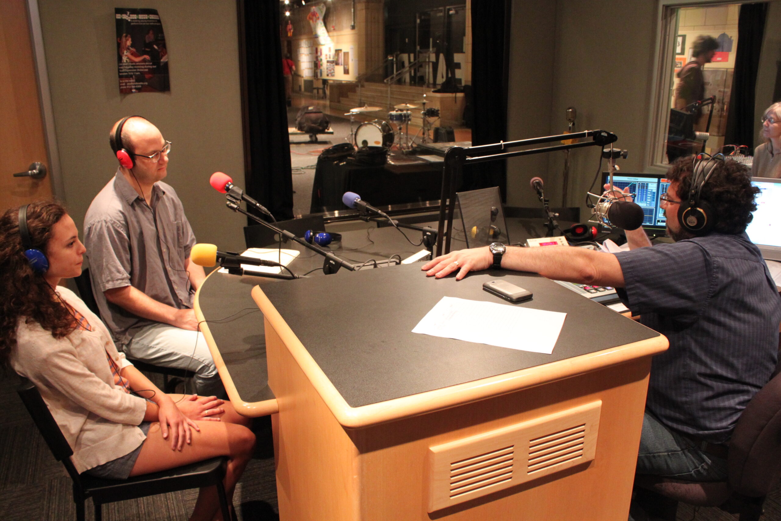 Two teens wearing headphones sitting behind microphones being interviewed by an adult wearing headphones speaking into a microphone in a radio studio