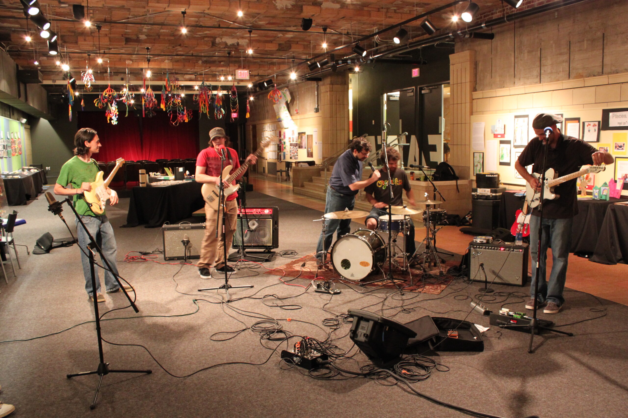 An adult holding an electric guitar, an adult playing an electric bass, and an adult speaking into a microphone leaning over an adult sitting at a drum set