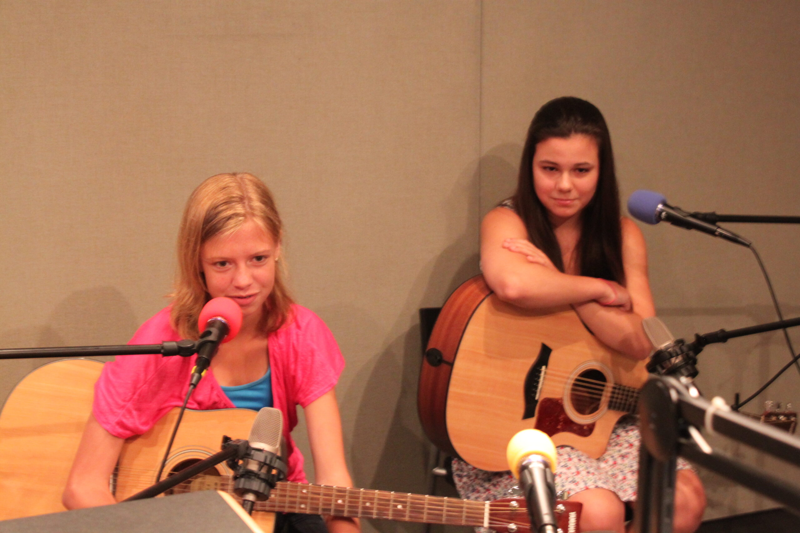 Two teens holding acoustic guitars sitting behind microphones in a radio studio