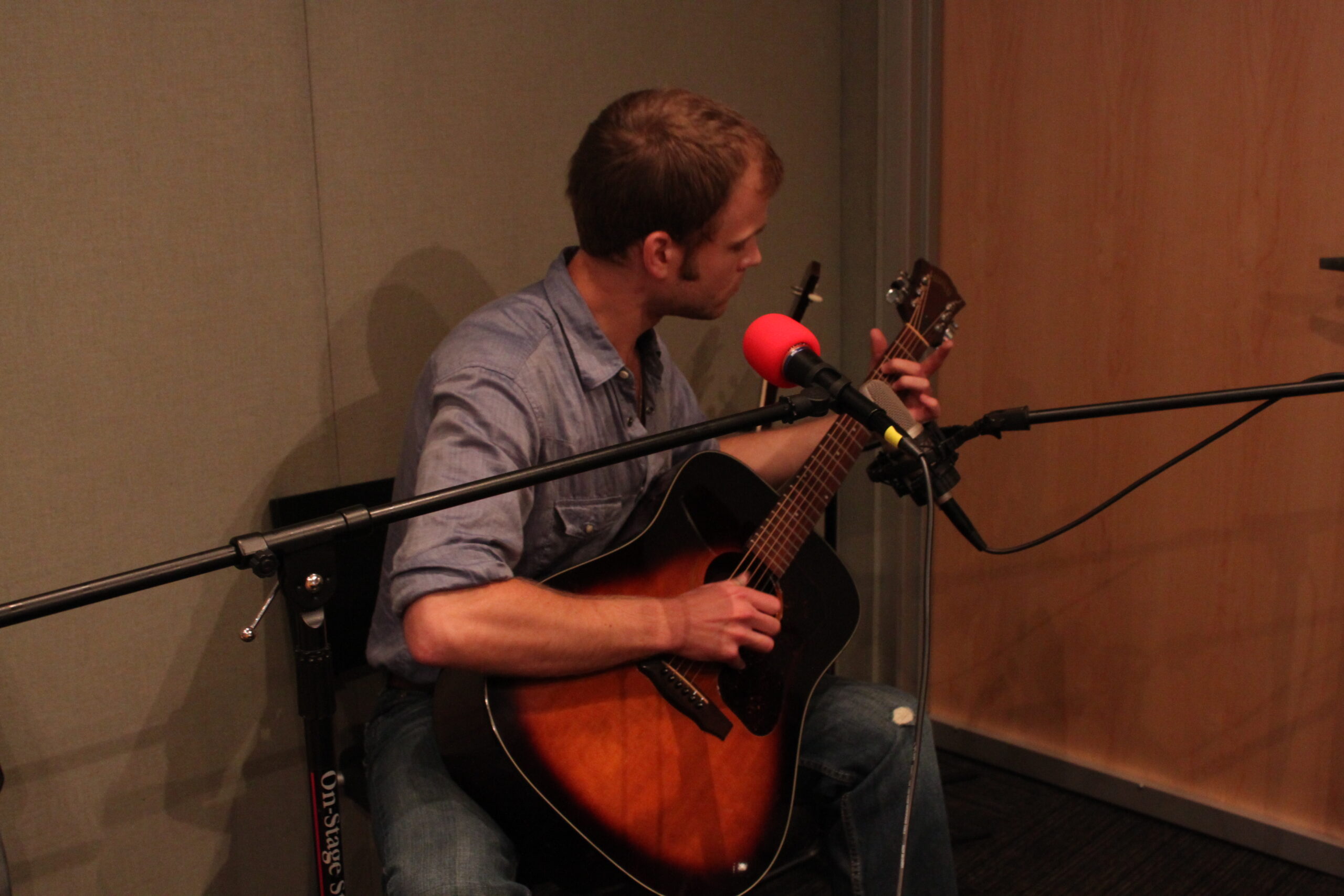 An adult playing acoustic guitar sitting behind a microphone in a radio studio