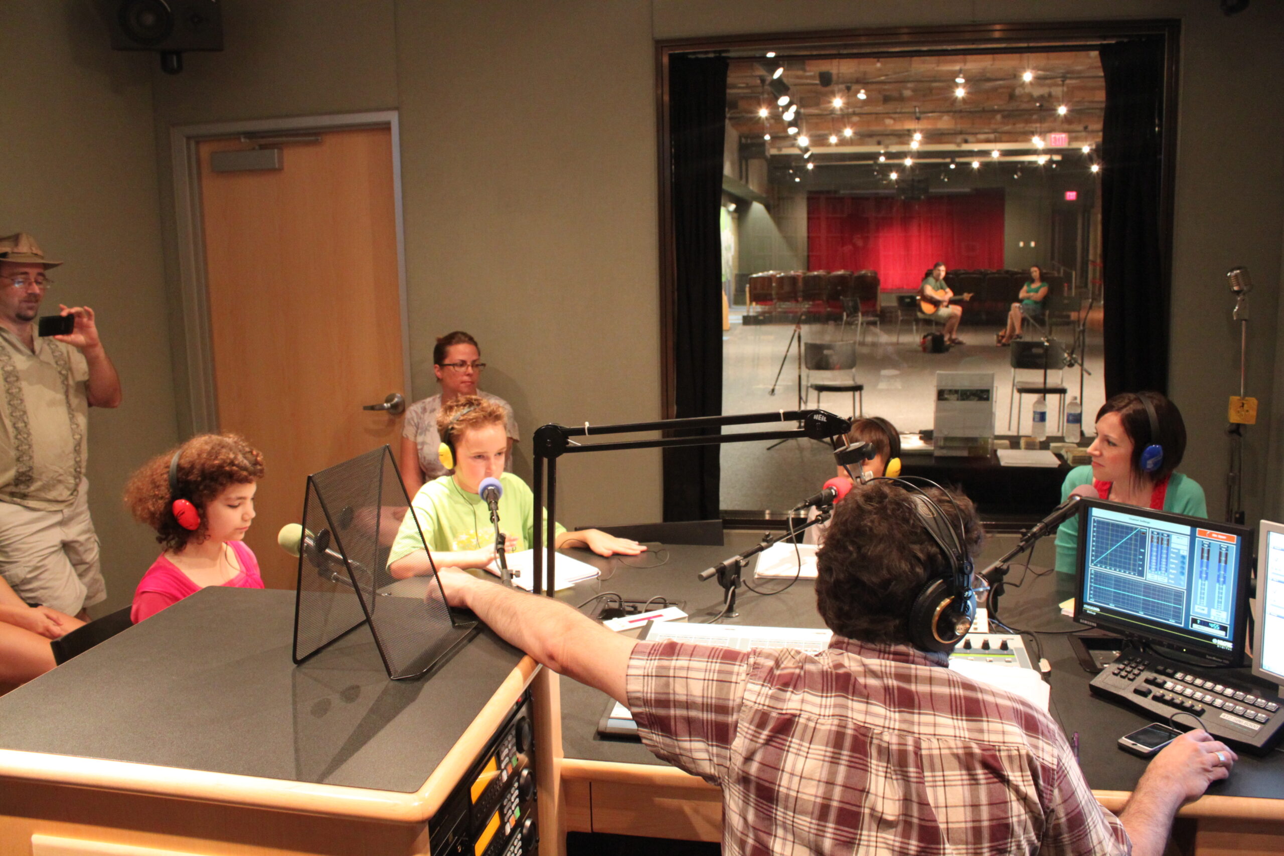 Four youth wearing headphones sitting behind microphones being interviewed by an adult wearing headphones sitting behind a microphone and an audio mixer as two adults in the background watch in a radio studio