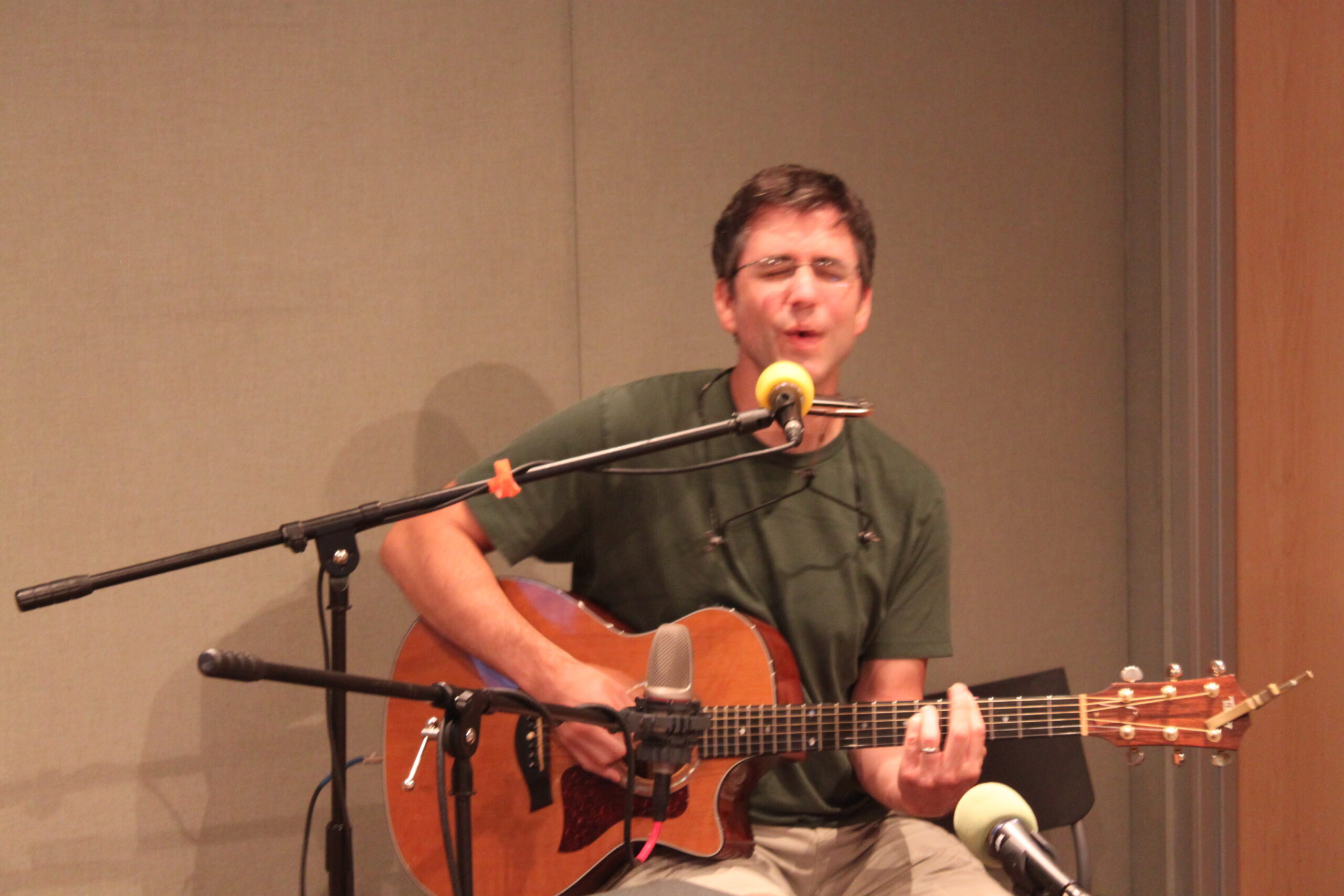 An adult playing acoustic guitar and singing into a microphone in a radio studio