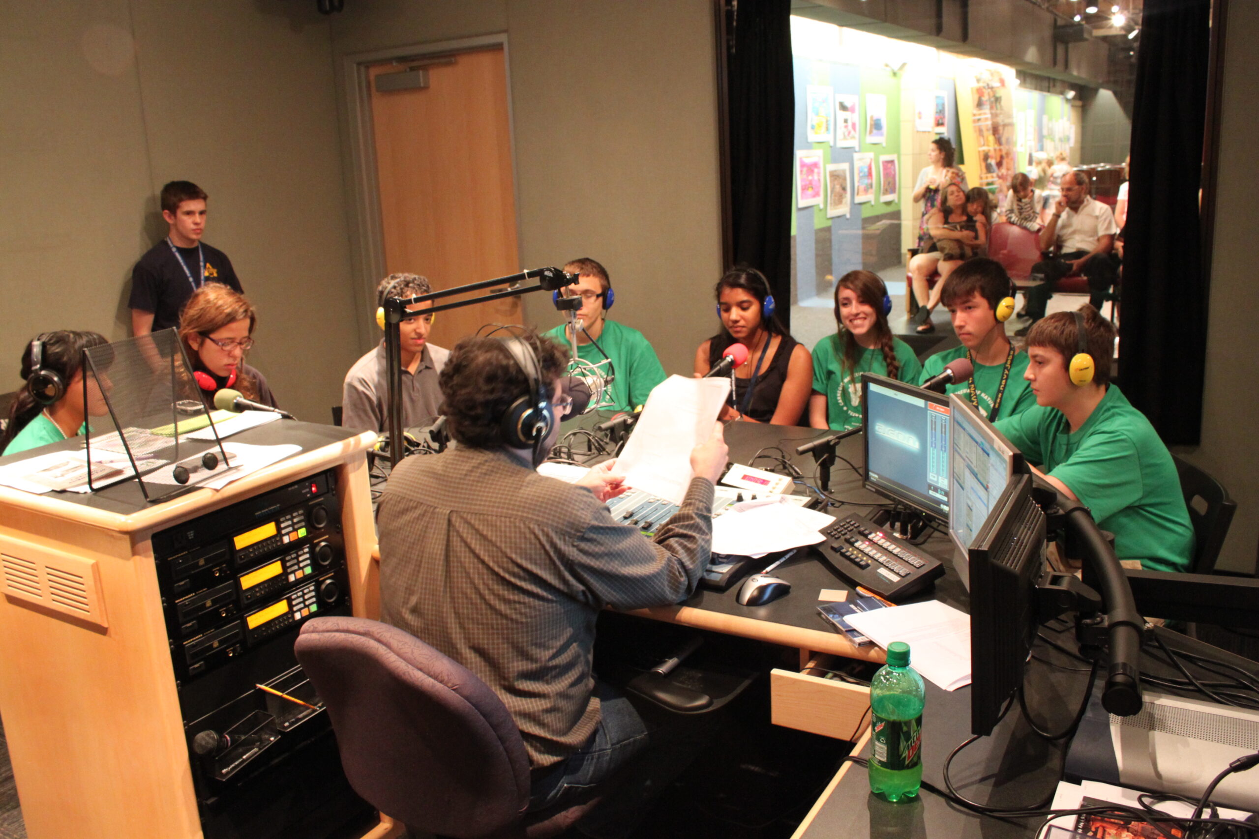 Eight teens wearing headphones sitting behind microphones being interviewed by an adult wearing headphones sitting behind an audio mixer and a microphone in a radio studio