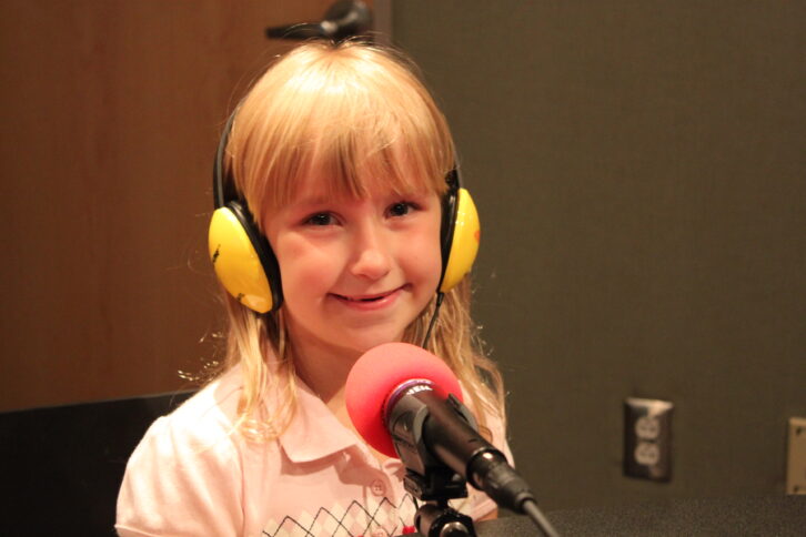 A youth wearing headphones smiling while sitting behind a microphone in a radio studio