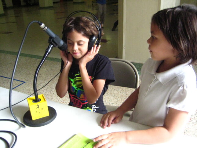 A youth looking at another youth holding headphones on their head while sitting behind a microphone