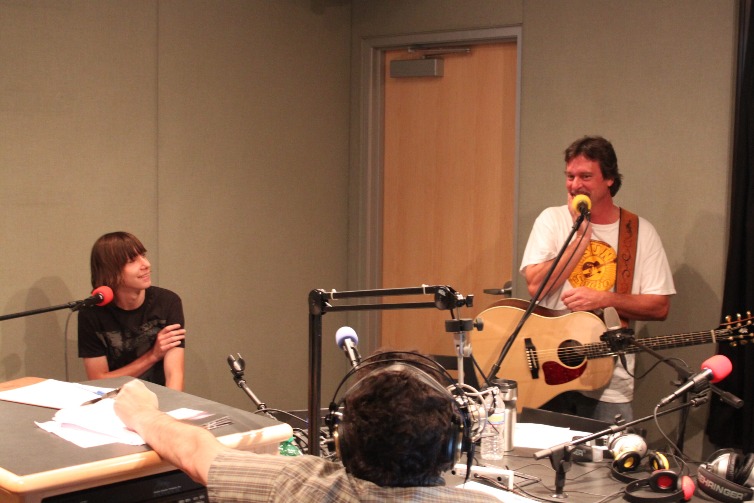 An adult holding an acoustic guitar and speaking into a microphone and a teen and an adult sitting behind microphones in a radio studio