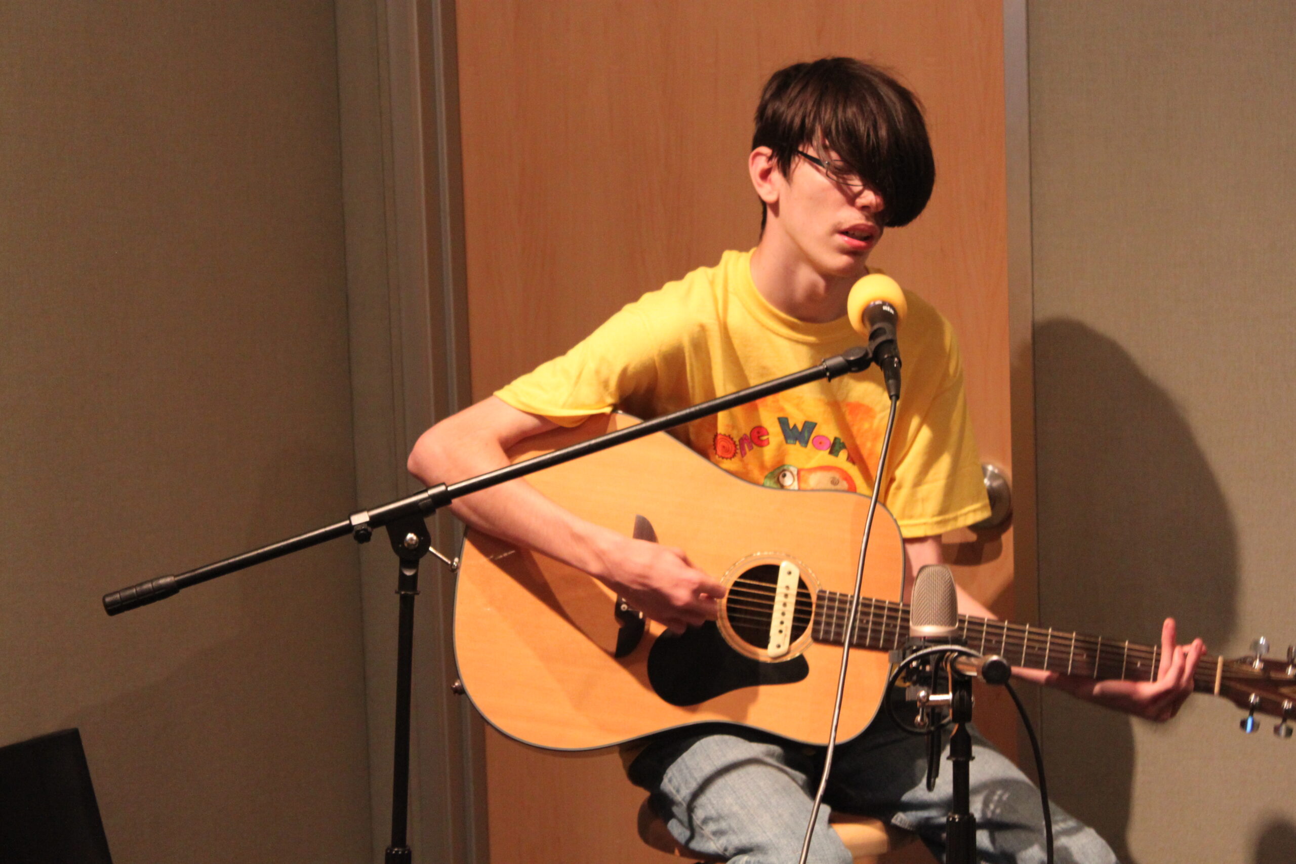 A teen playing an acoustic guitar and singing into a microphone in a radio studio