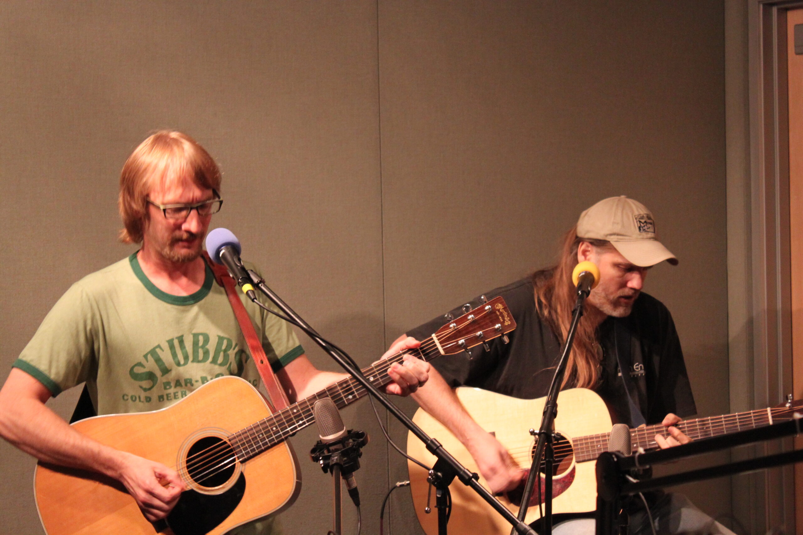 Two adults playing acoustic guitars standing behind microphones in a radio studio