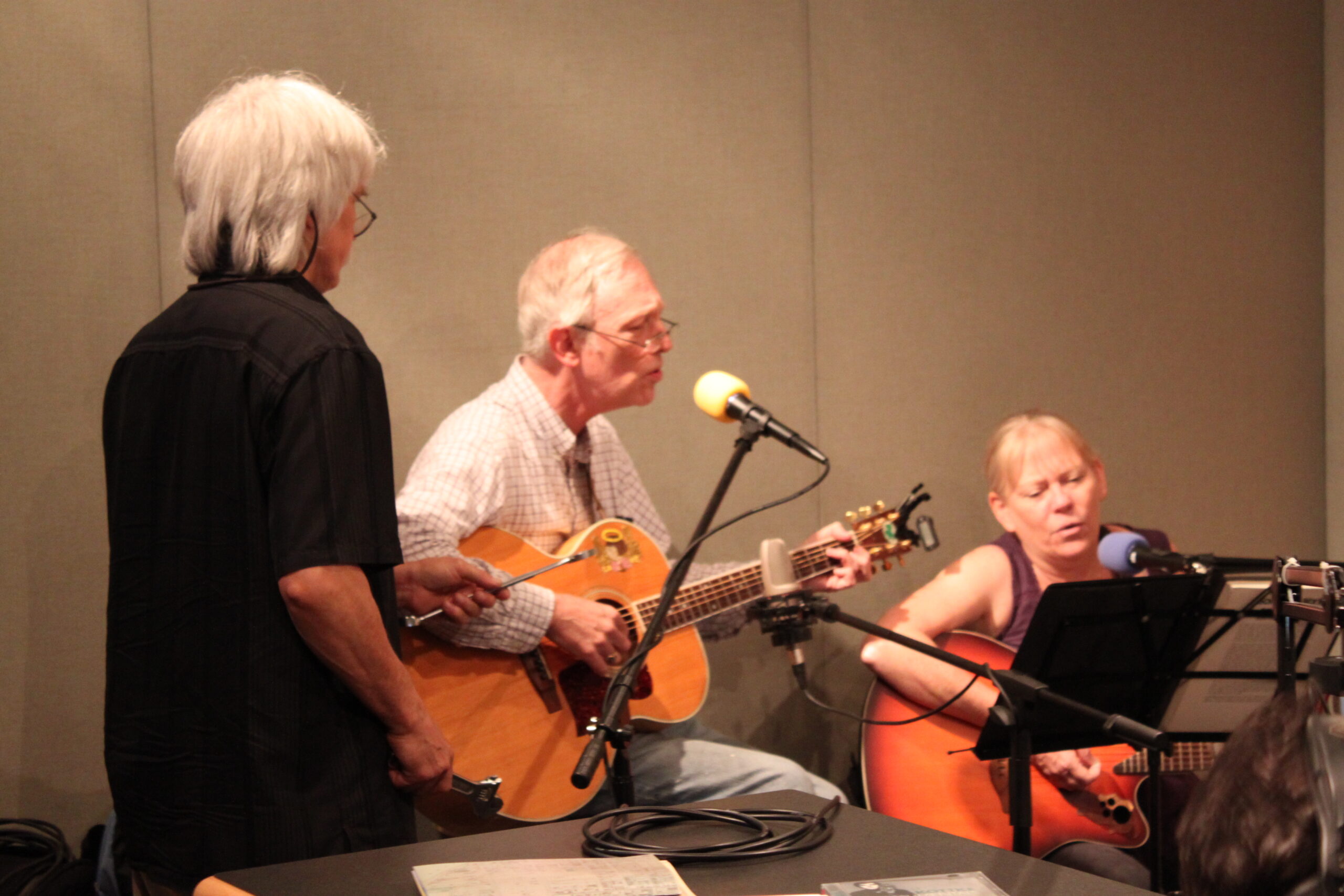 Two adults playing acoustic guitars and singing into microphones and an adult watching in a radio studio