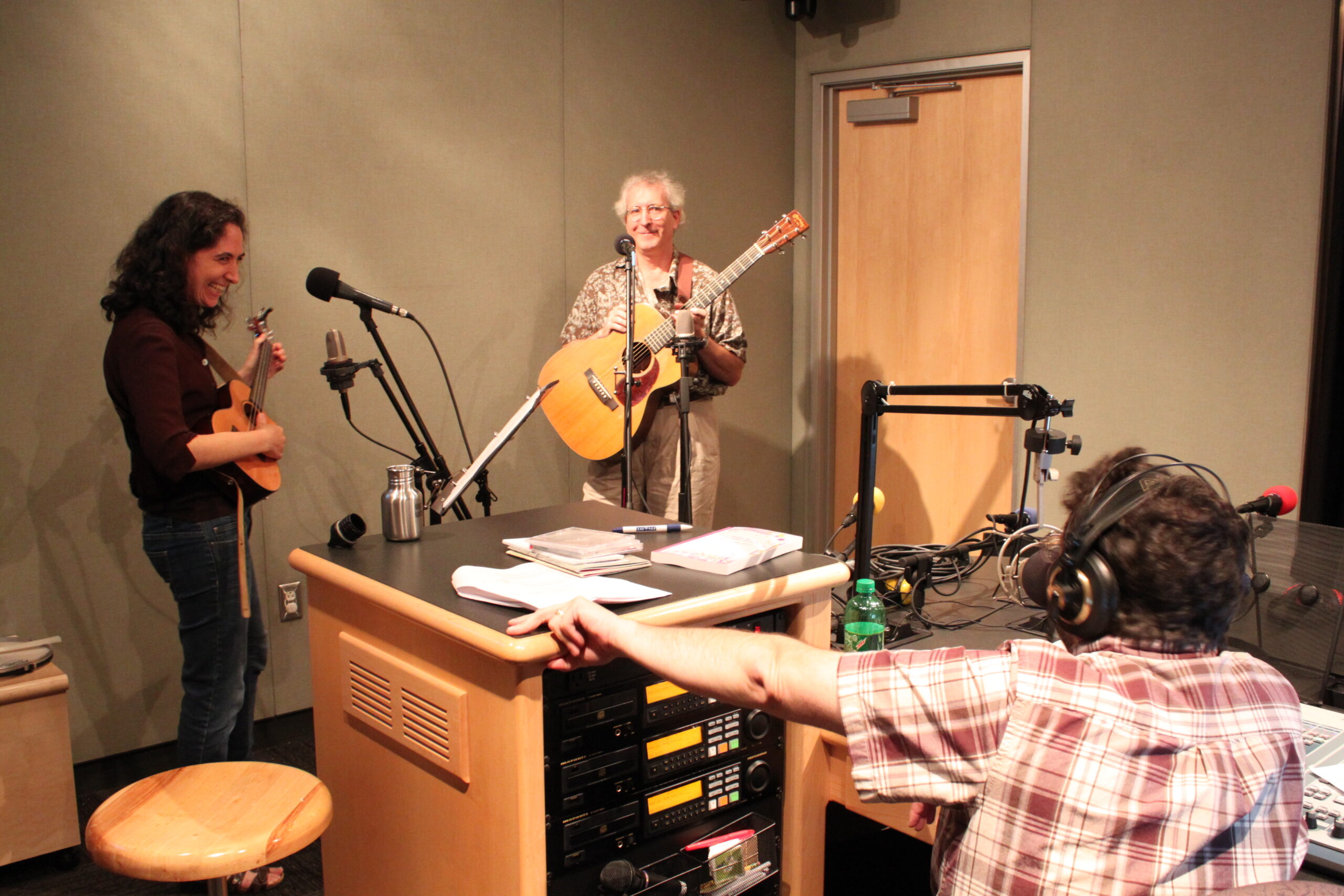 An adult playing a ukulele and an adult playing an acoustic guitar, both singing into microphones as an adult wearing headphones sitting behind a microphone and an audio mixer listens in a radio studio