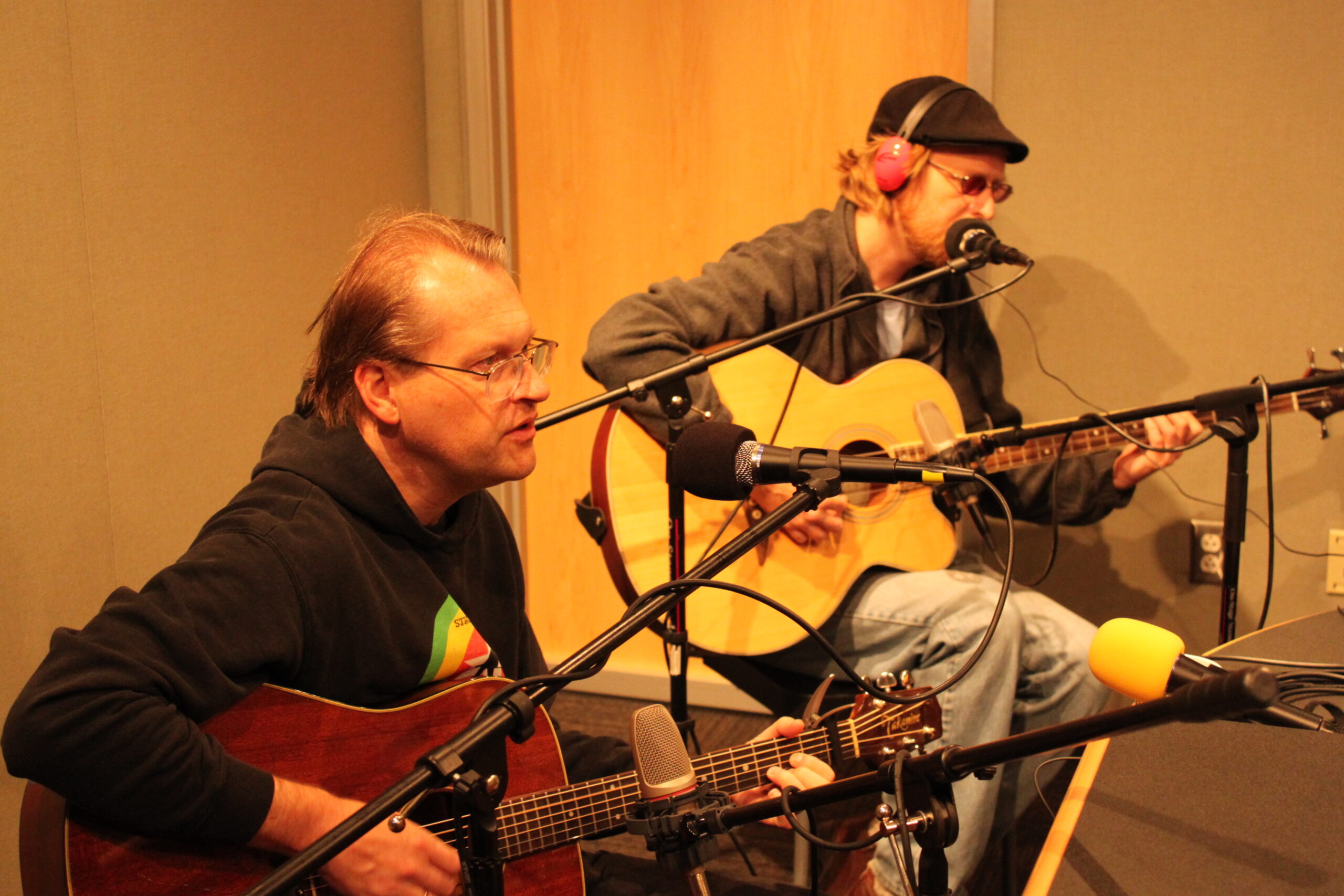 Two adults playing acoustic guitars in a radio studio