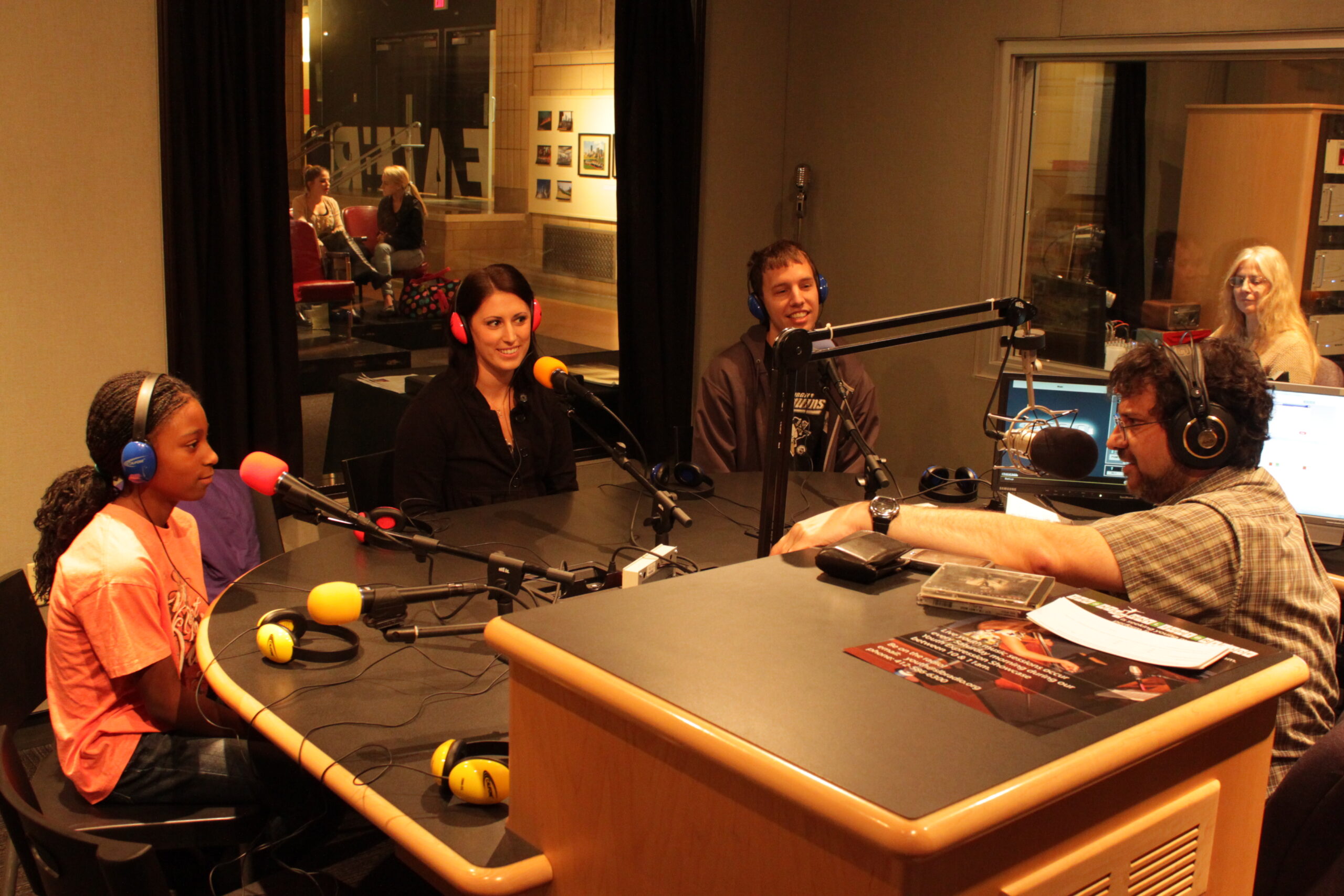 A youth and two adults wearing headphones speaking into microphones being interviewed by an adult wearing headphones sitting behind a microphone in a radio studio