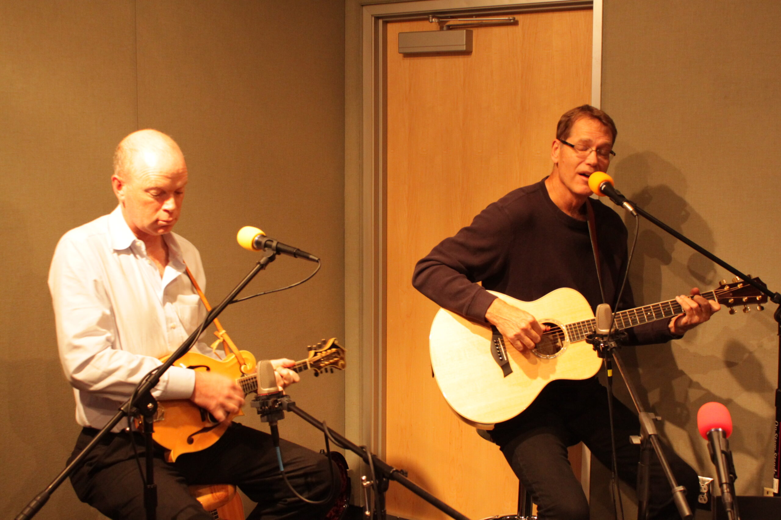 Two adults playing acoustic guitars and singing into microphones in a radio studio