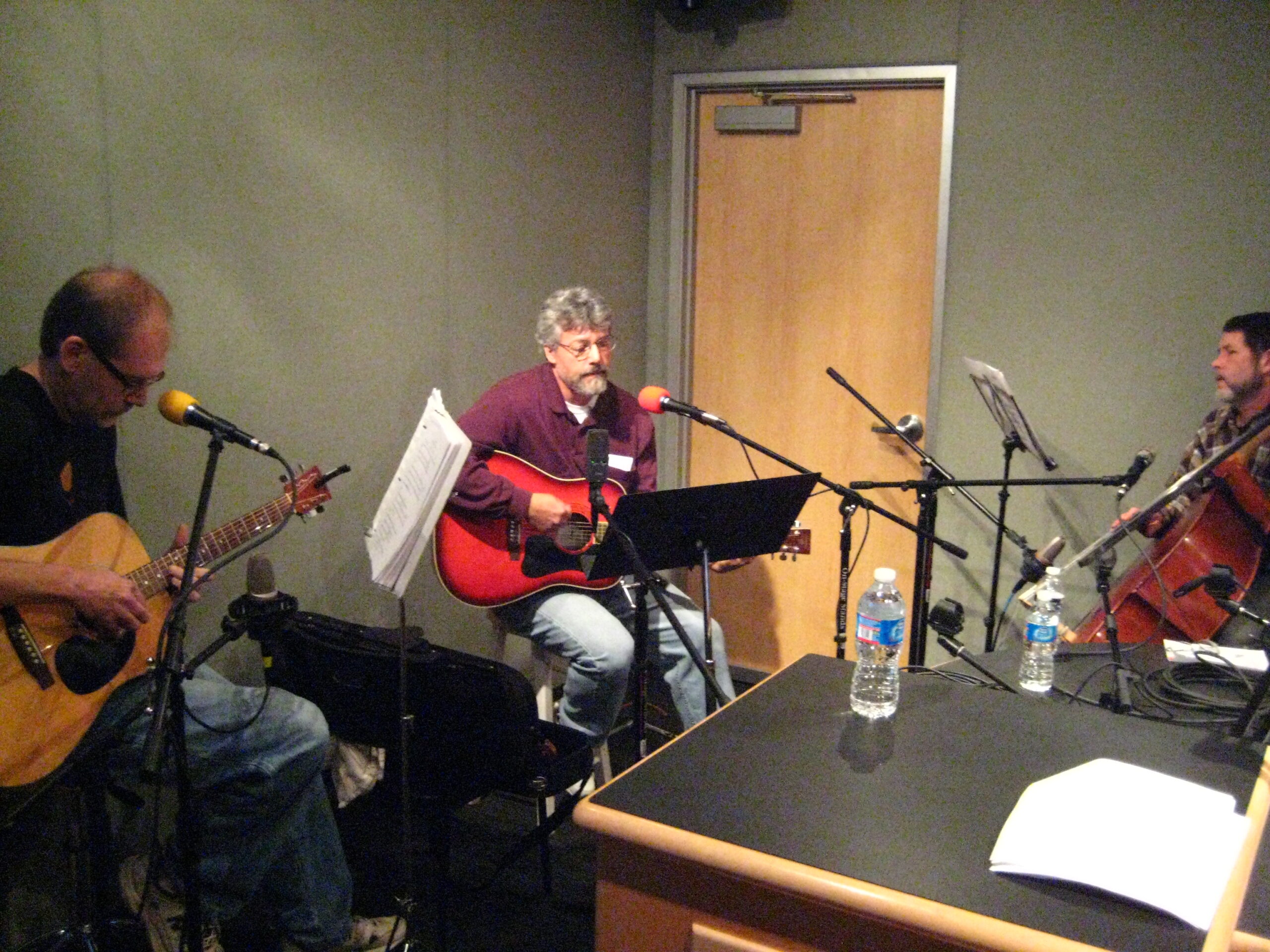 Two adults playing acoustic guitars and an adult playing an upright bass in a radio studio