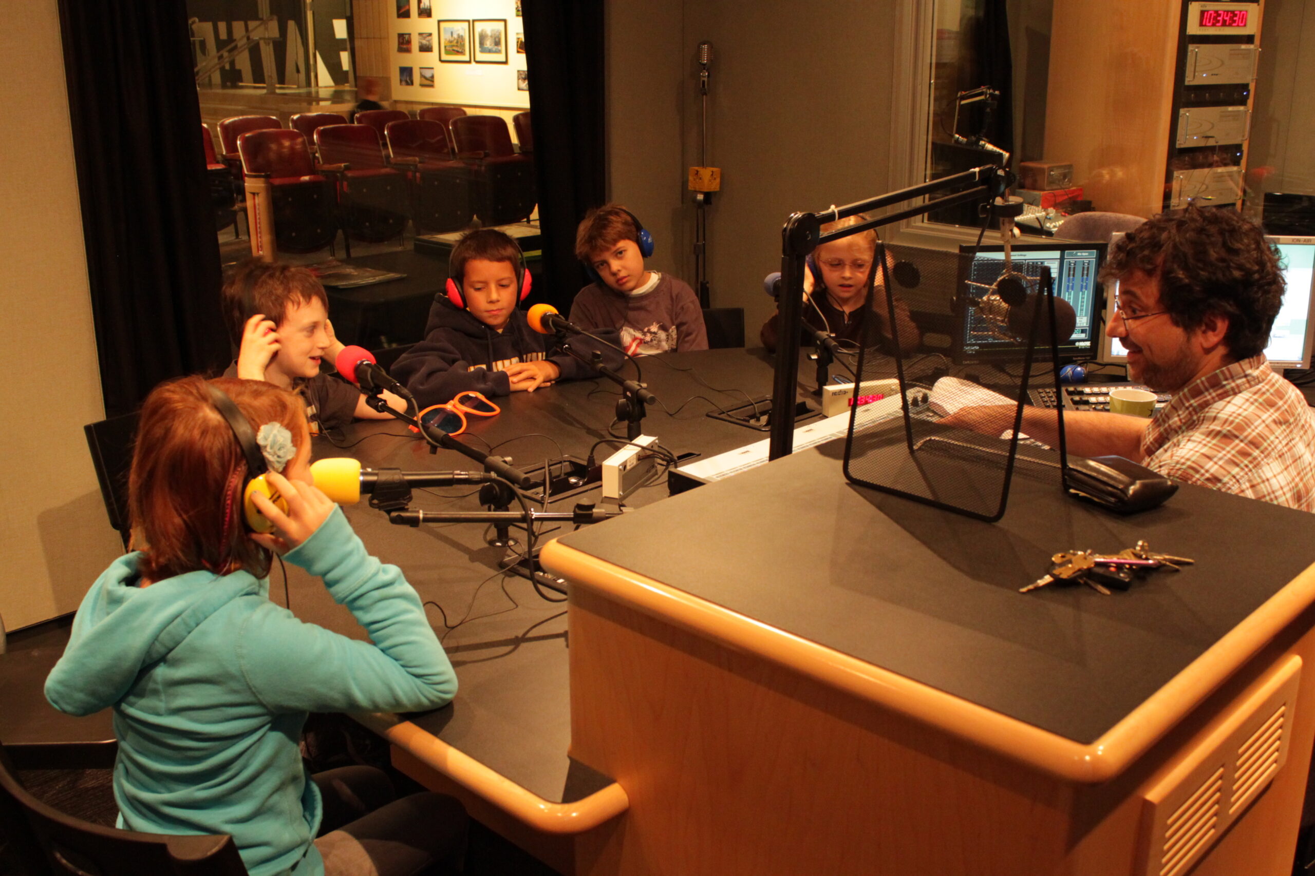 Five youth wearing headphones speaking into microphones being interviewed by an adult sitting behind a microphone in a radio studio