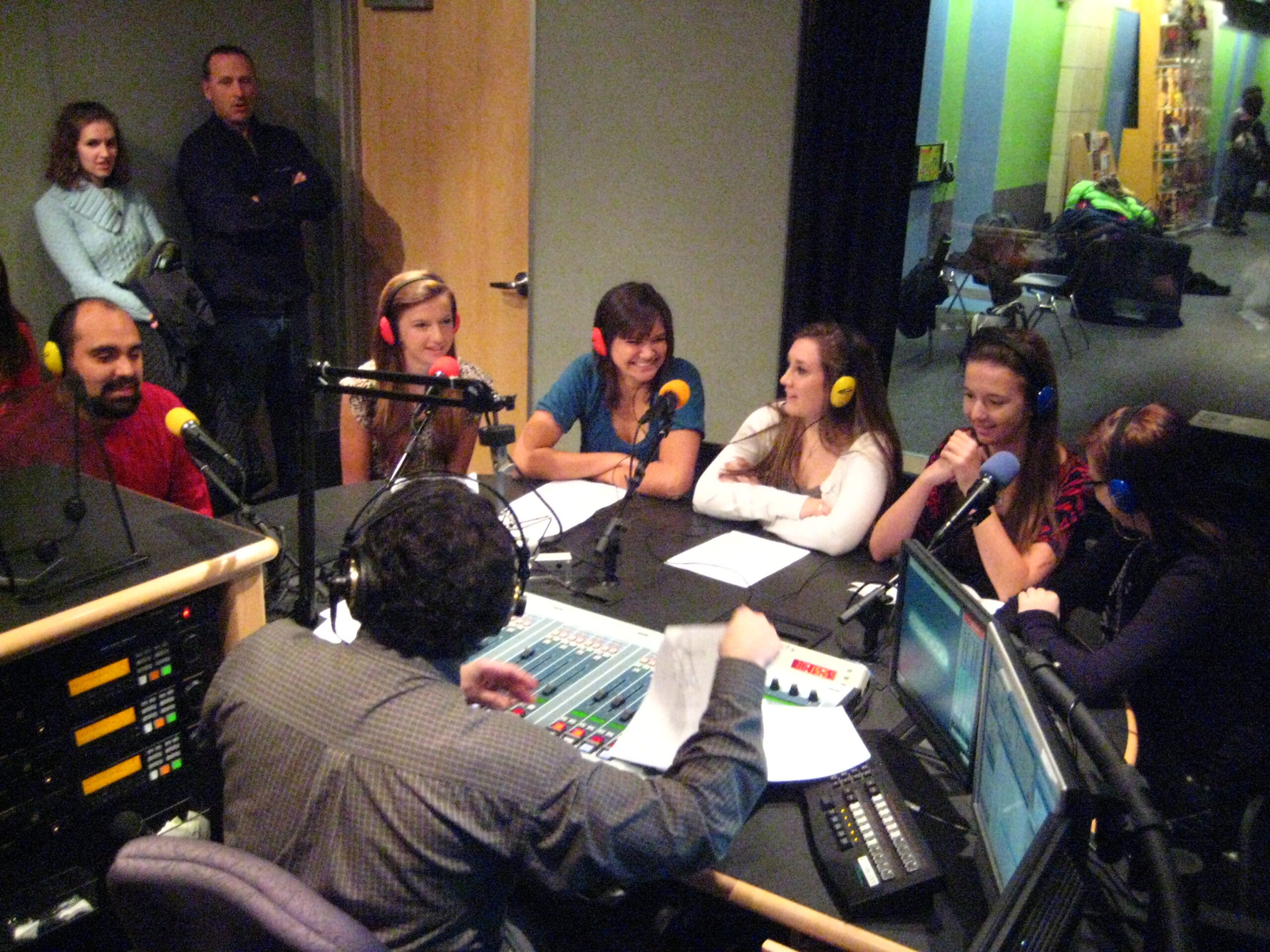 Five teens and an adult wearing headphones speaking into microphones being interviewed by an adult wearing headphones sitting behind a microphone and an audio mixer in a radio studio