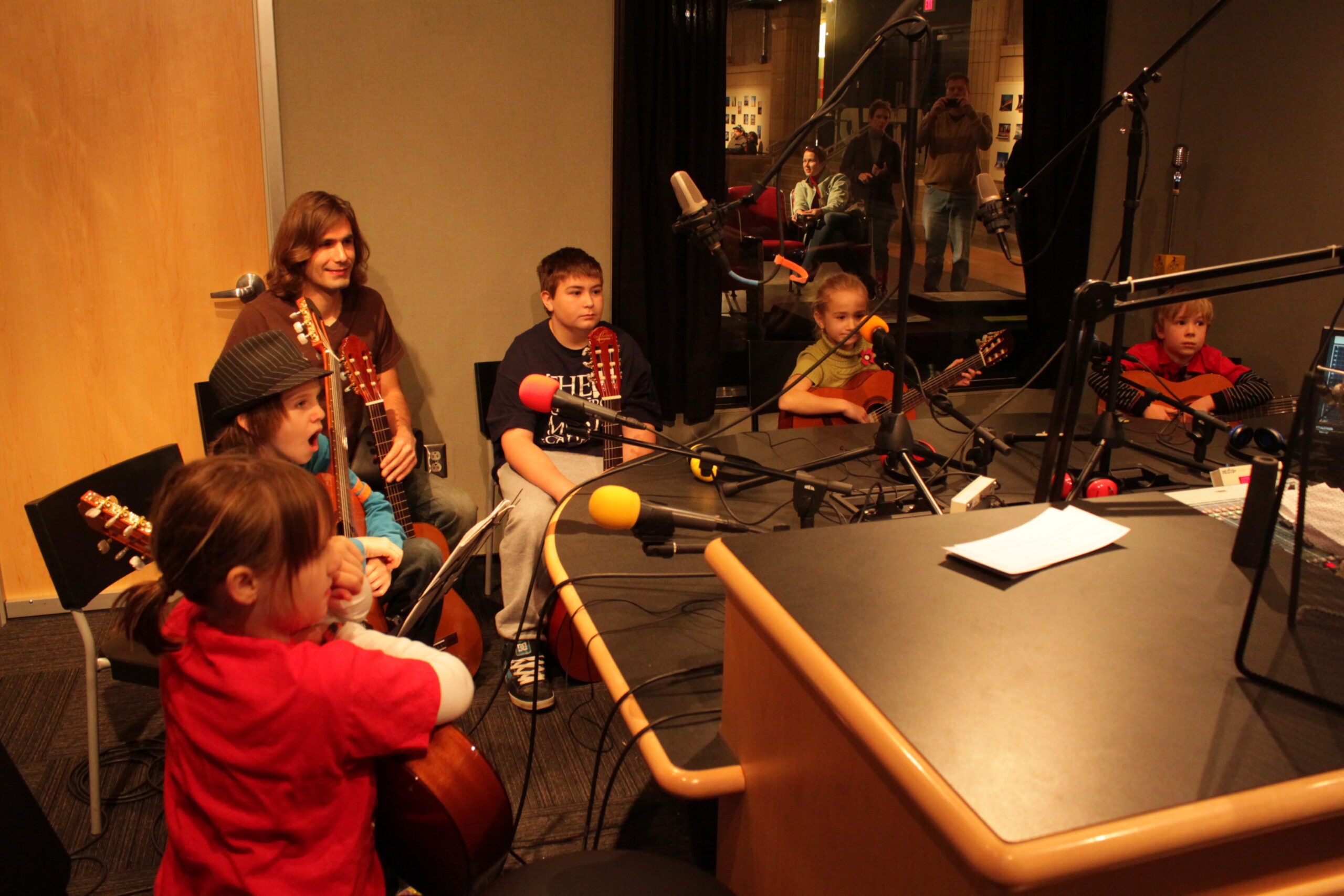 Five youth and one adult holding acoustic guitars in a radio studio