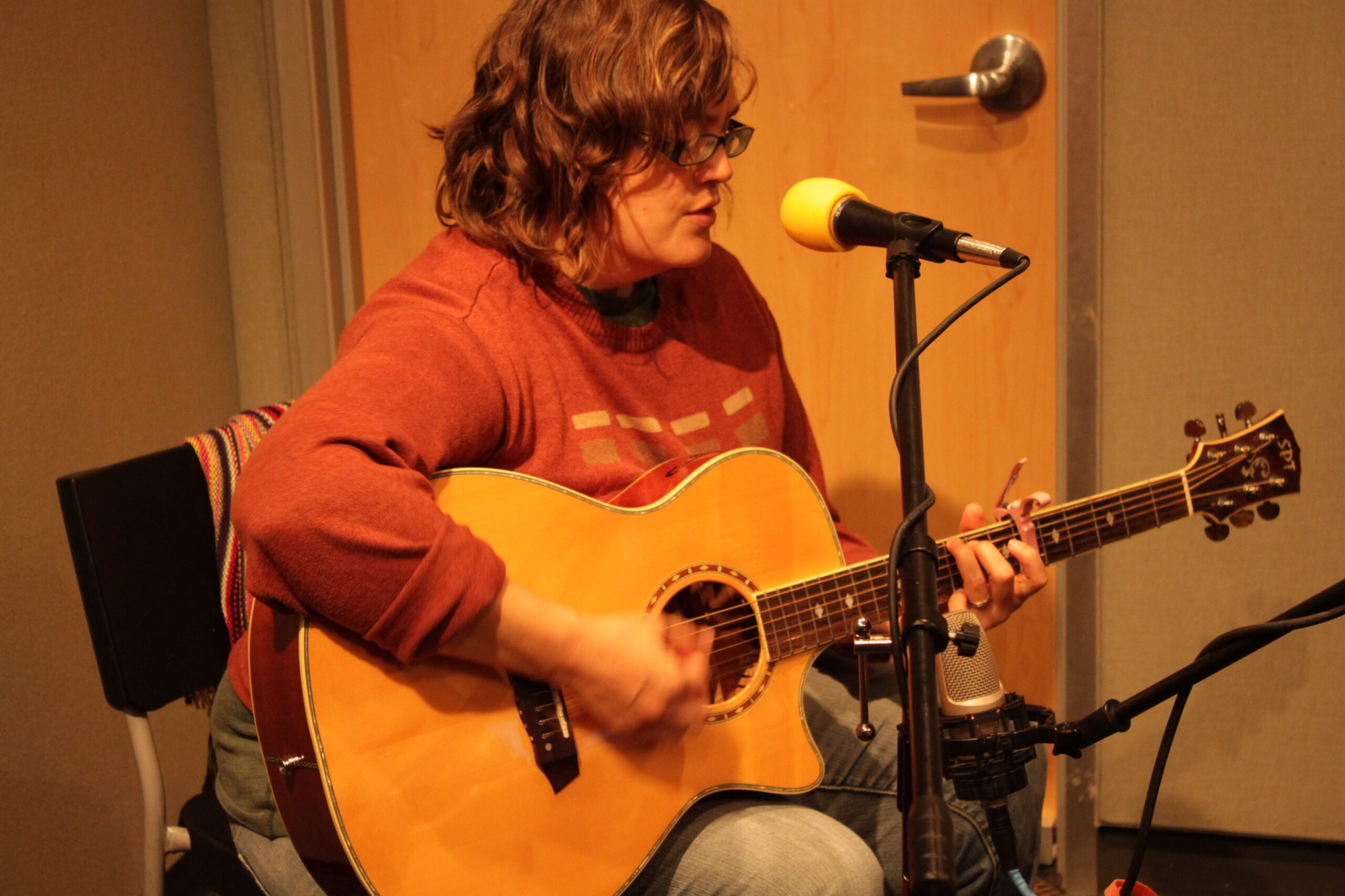 An adult playing an acoustic guitar sitting behind a microphone in a radio studio