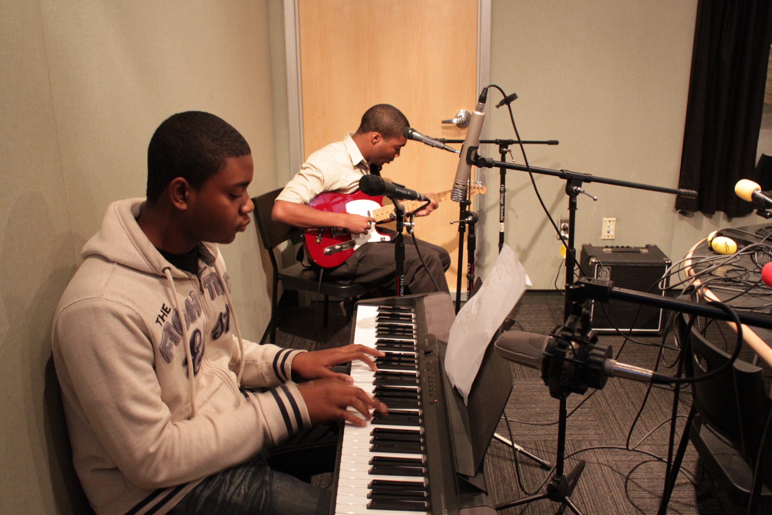 A teen playing a piano keyboard and a teen playing electric bass in a radio studio