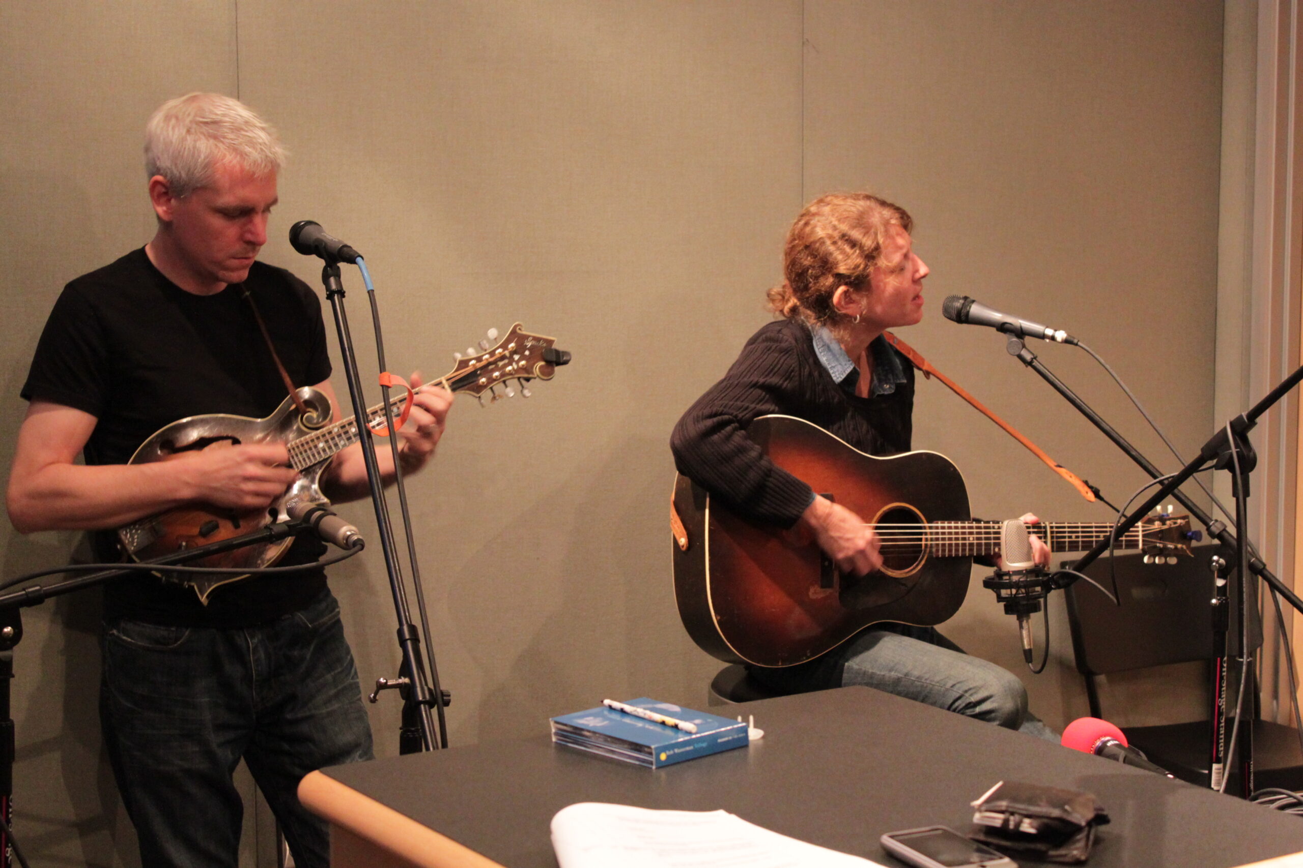 An adult playing mandolin and an adult playing acoustic guitar and singing into a microphone in a radio studio