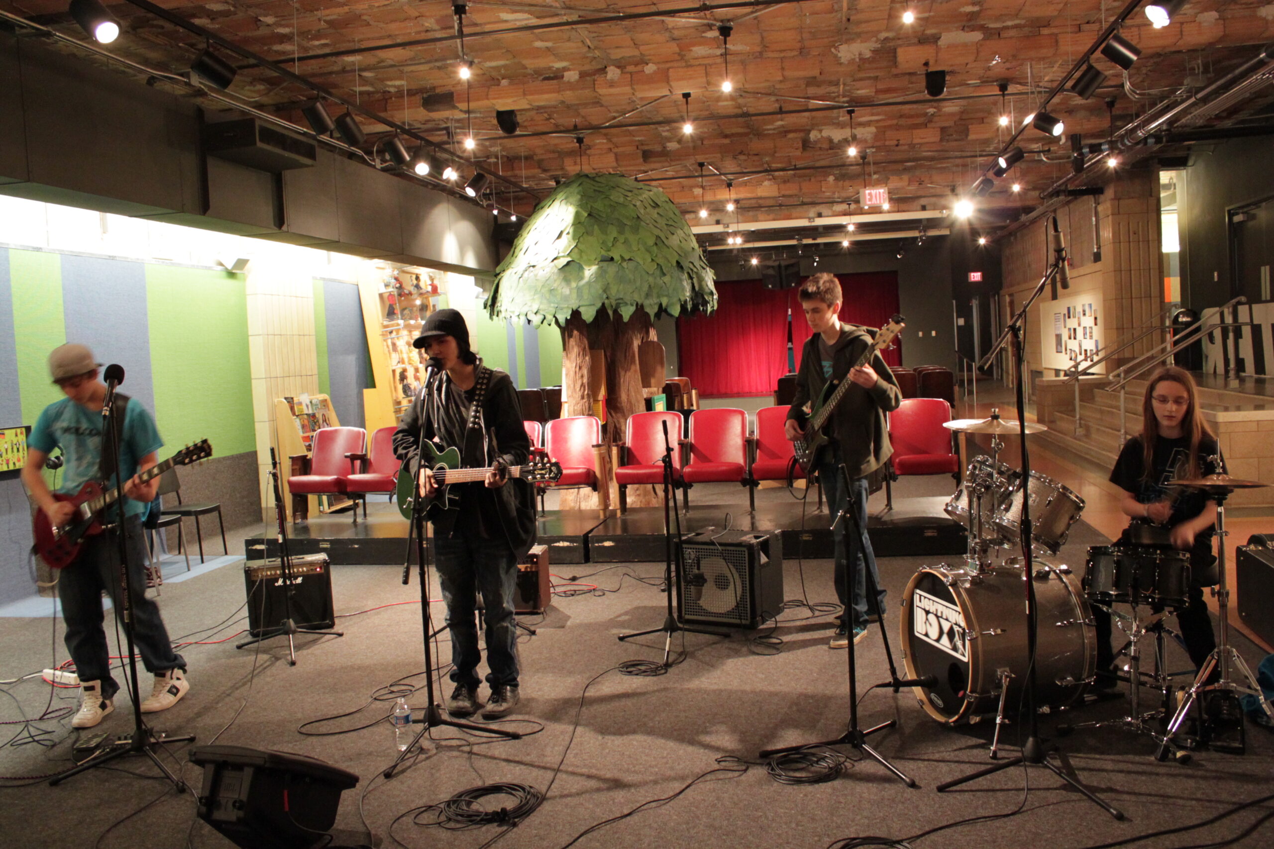 A teen playing electric guitar, a teen playing electric guitar and singing into a microphone, a teen playing electric bass, and a teen playing a drum set.
