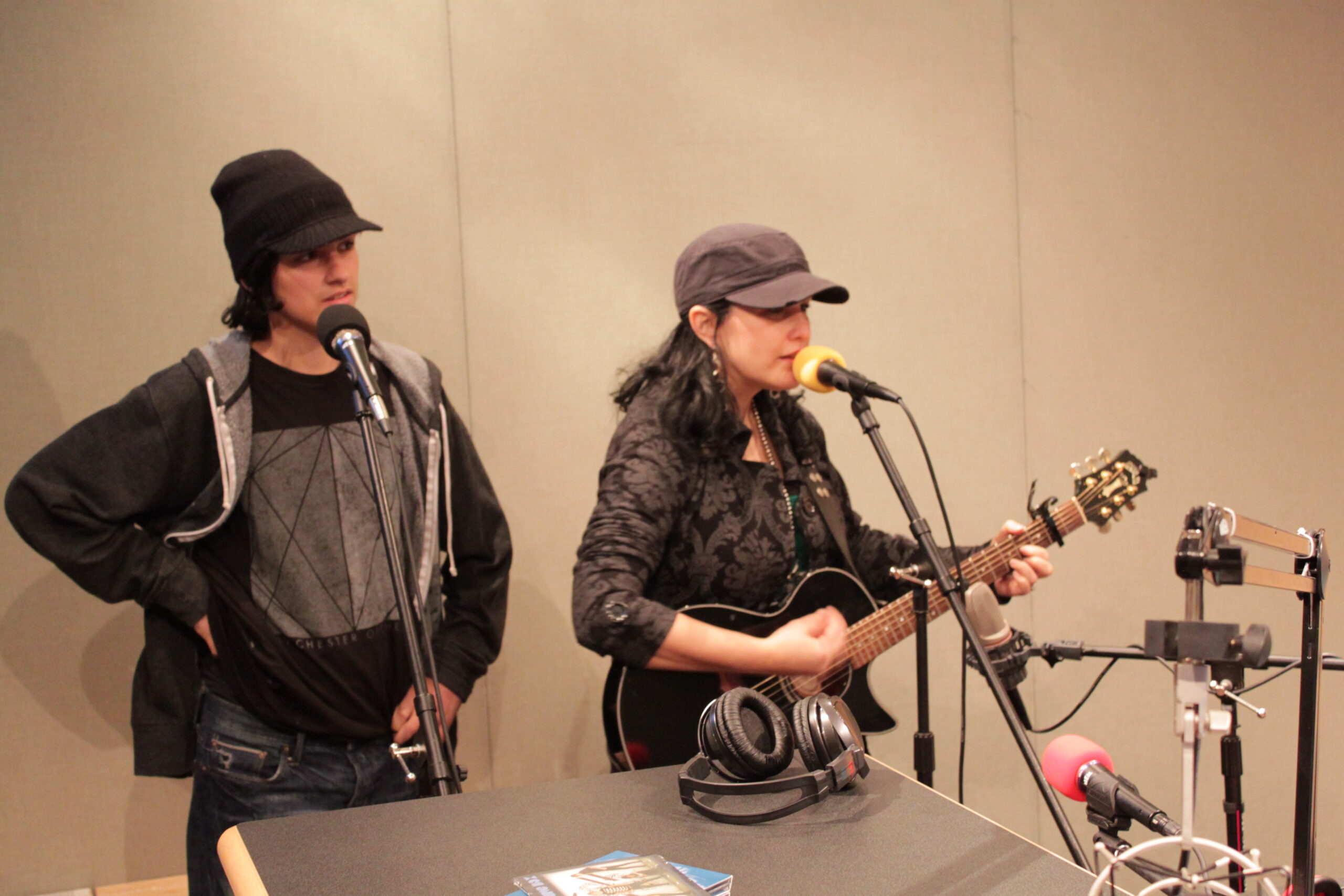 An adult singing into a microphone and an adult playing an acoustic guitar and singing into a microphone in a radio studio