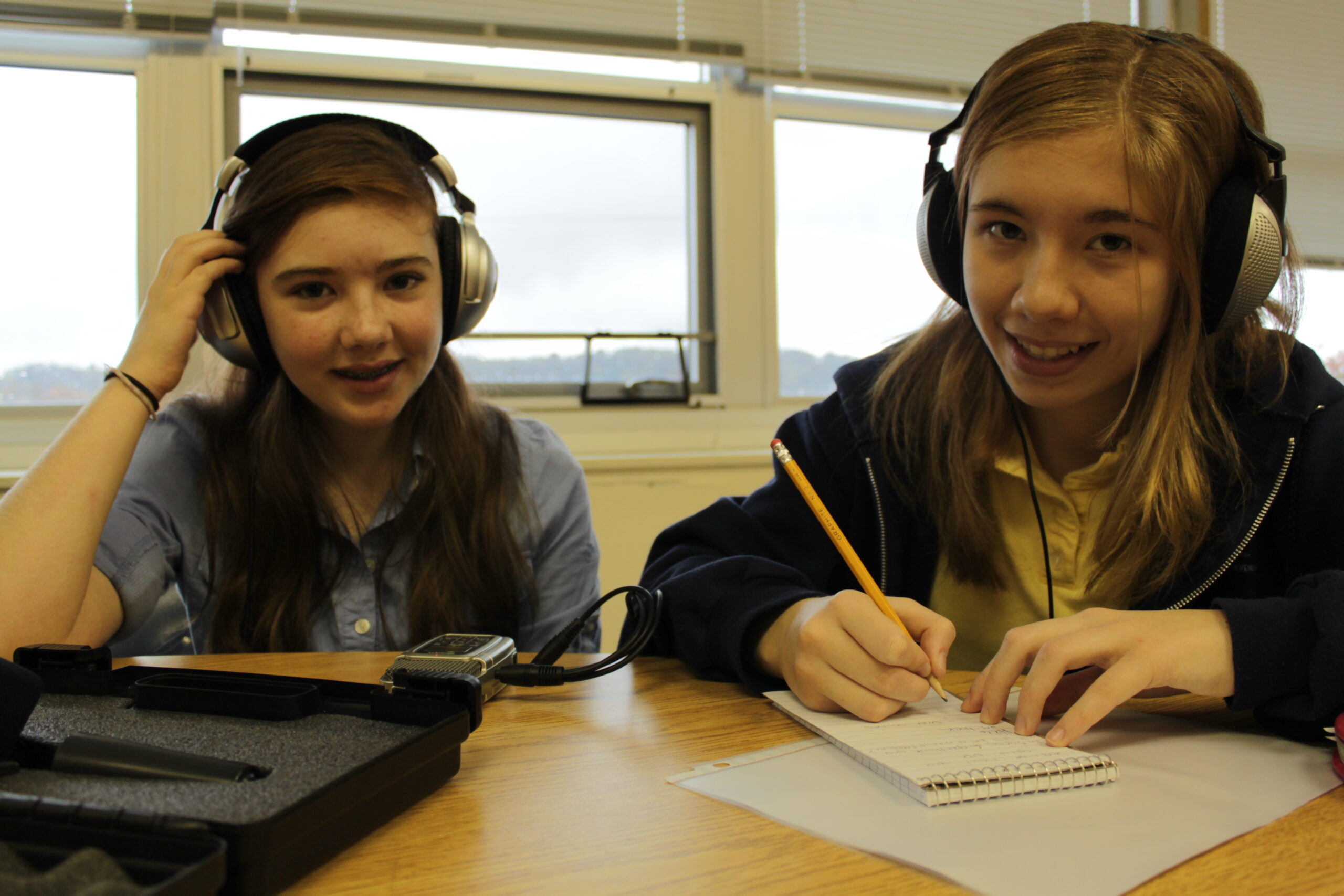 Two youth wearing headphones sitting at a classroom table, one writing on a piece of paper