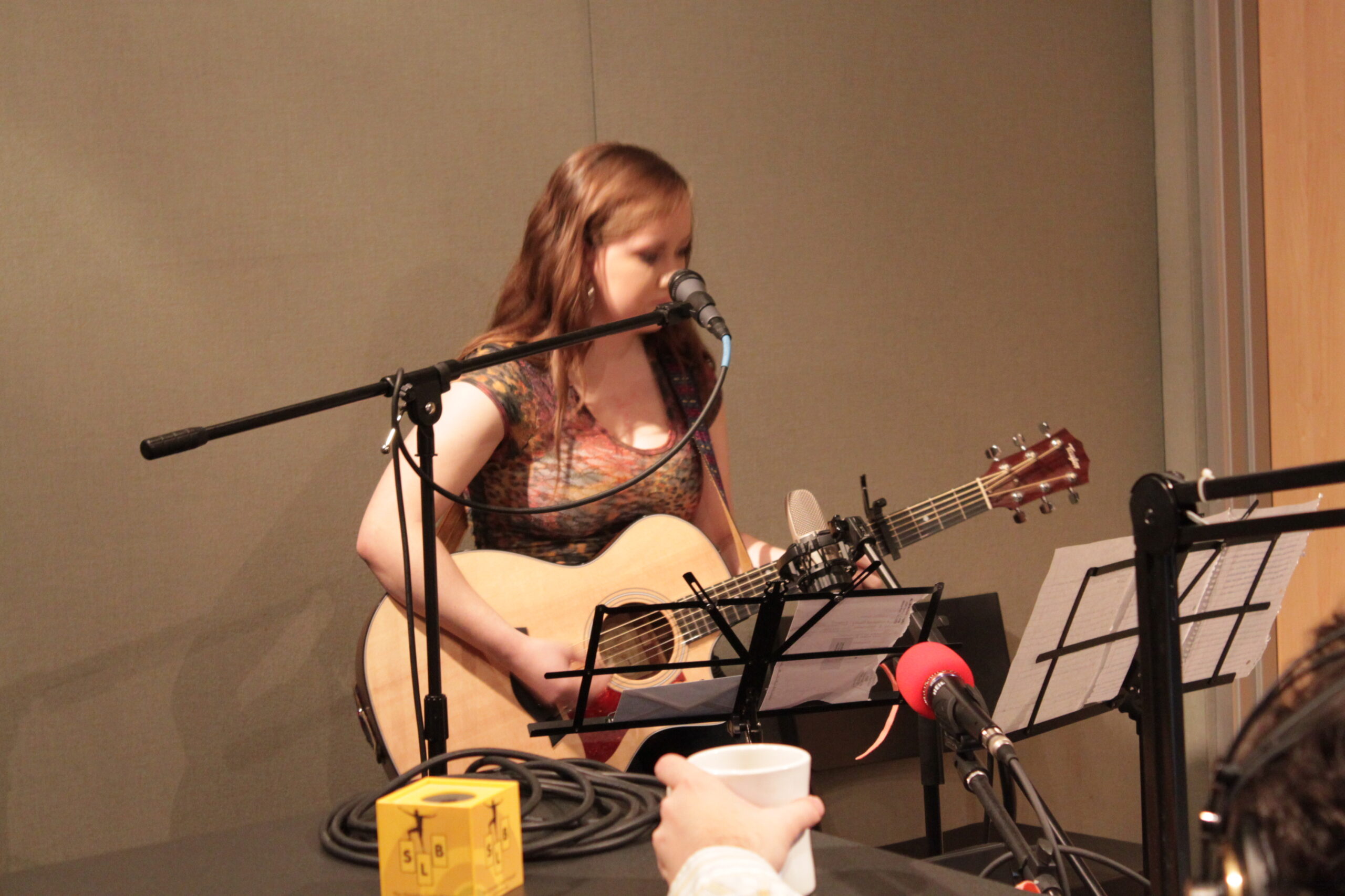 A teen playing acoustic guitar sitting behind a microphone and music stands in a radio studio