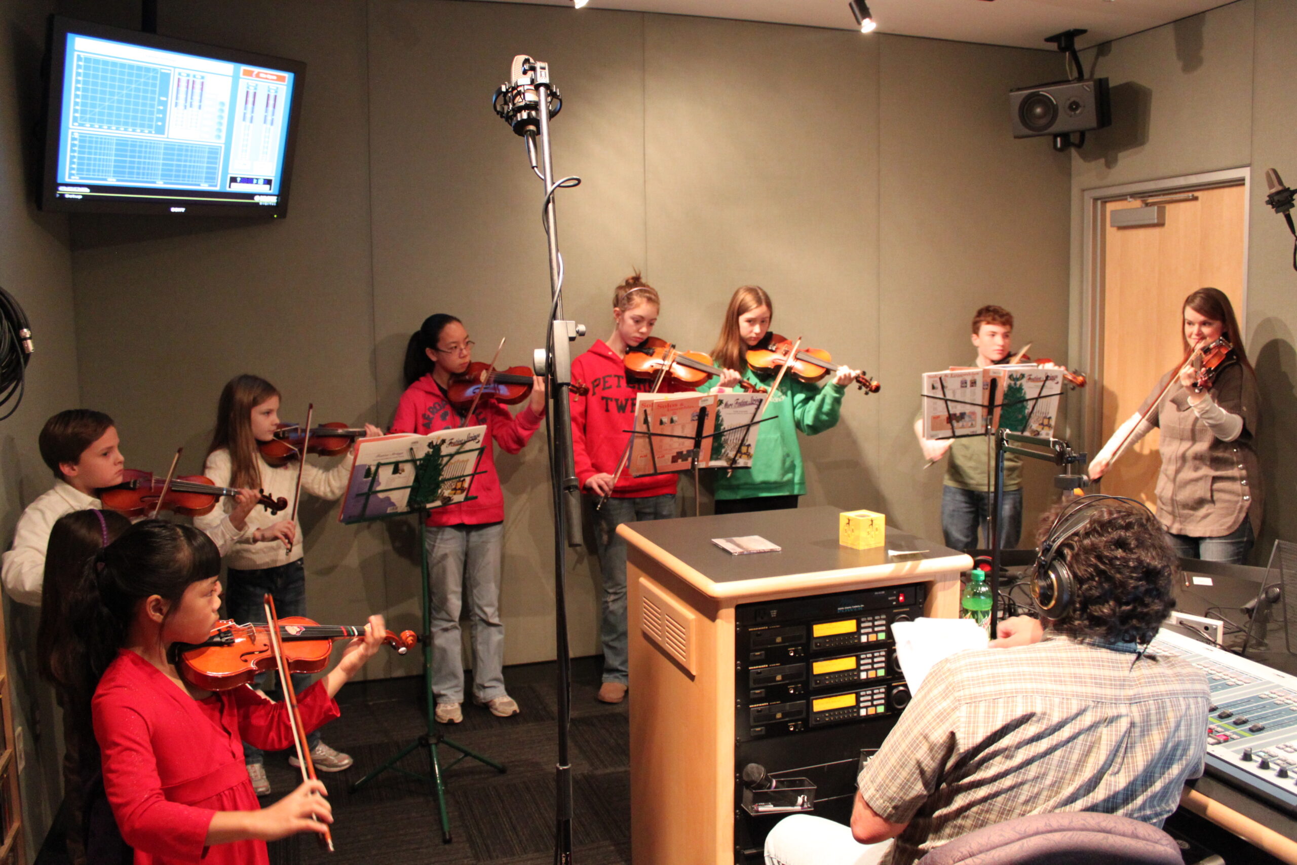 Eight youth and an adult playing violins and looking at sheet music on music stands in a radio studio