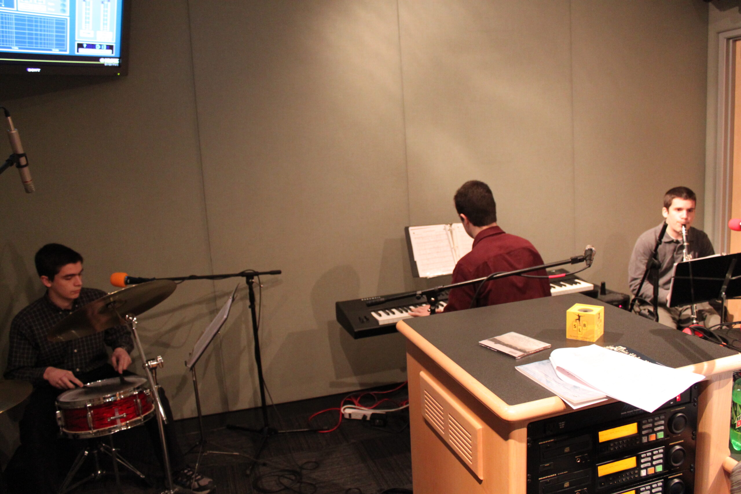 A teen playing a drum set, a teen playing a piano keyboard, and a teen playing a clarinet in a radio studio
