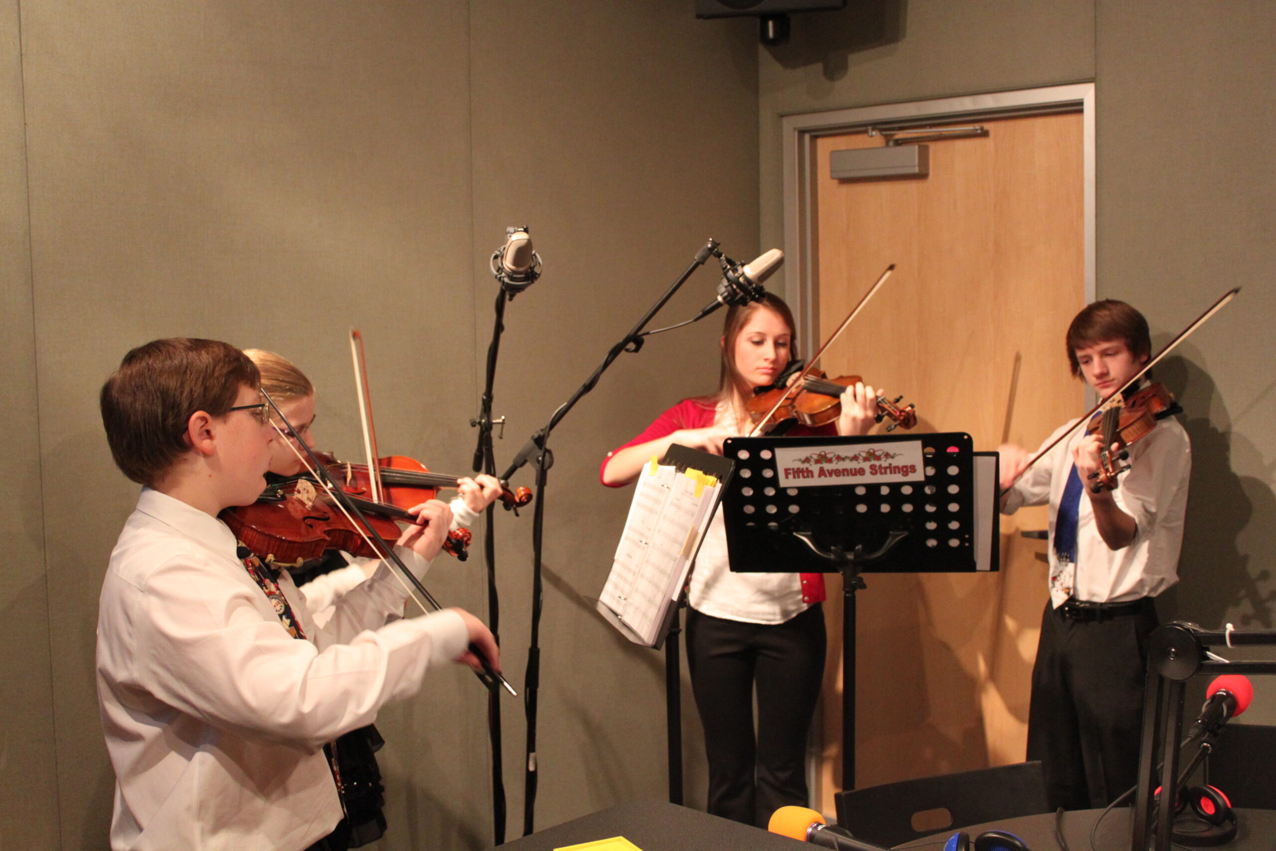 Four youth playing violins looking at sheet music on music stands in a radio studio