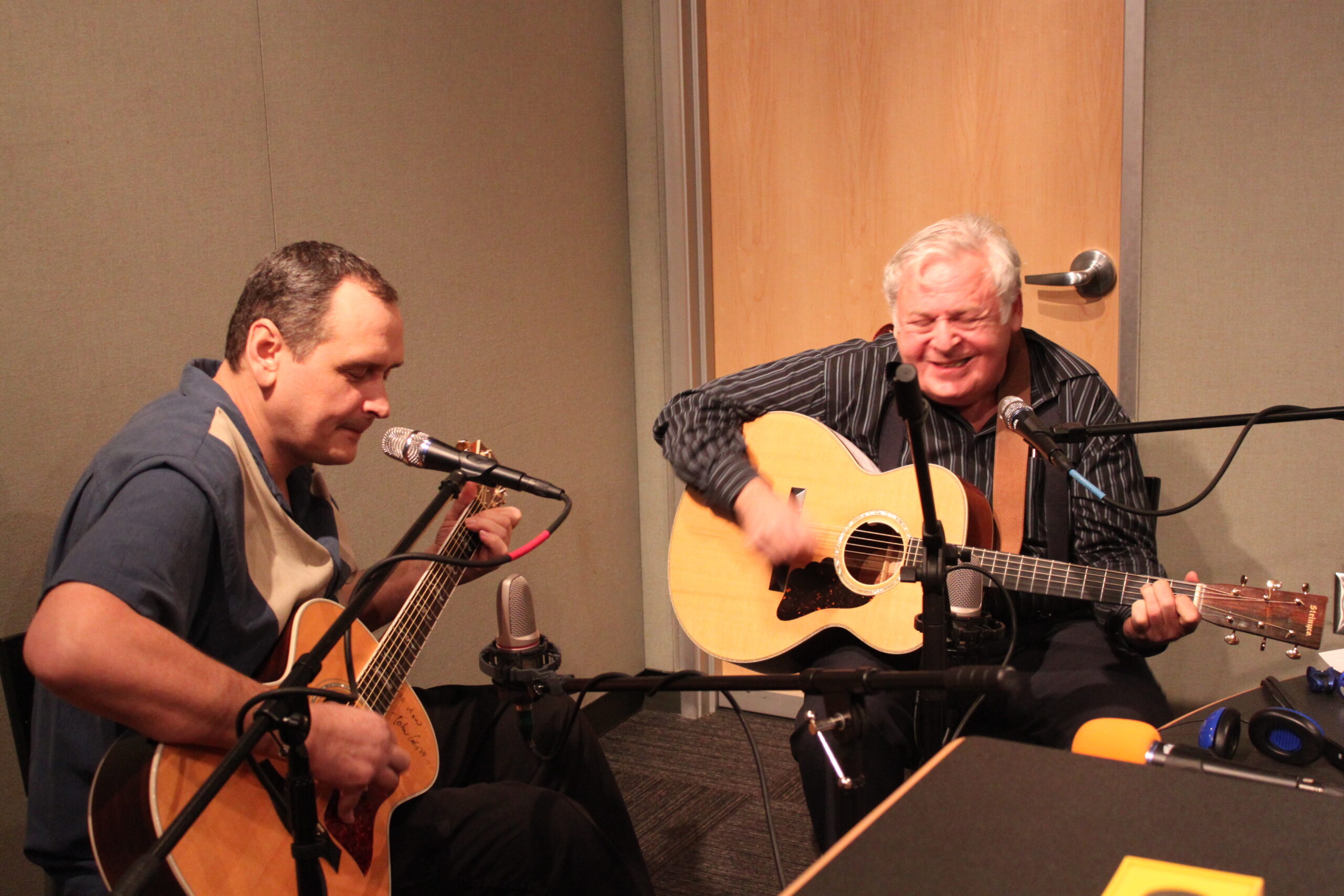 Two adults sitting behind microphones holding acoustic guitars in a radio studio