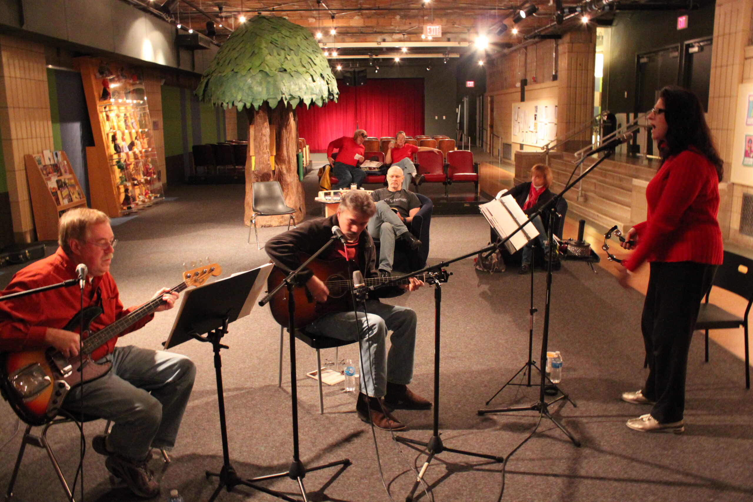 An adult playing an electric bass, an adult playing an electric guitar, and an adult singing into a microphone
