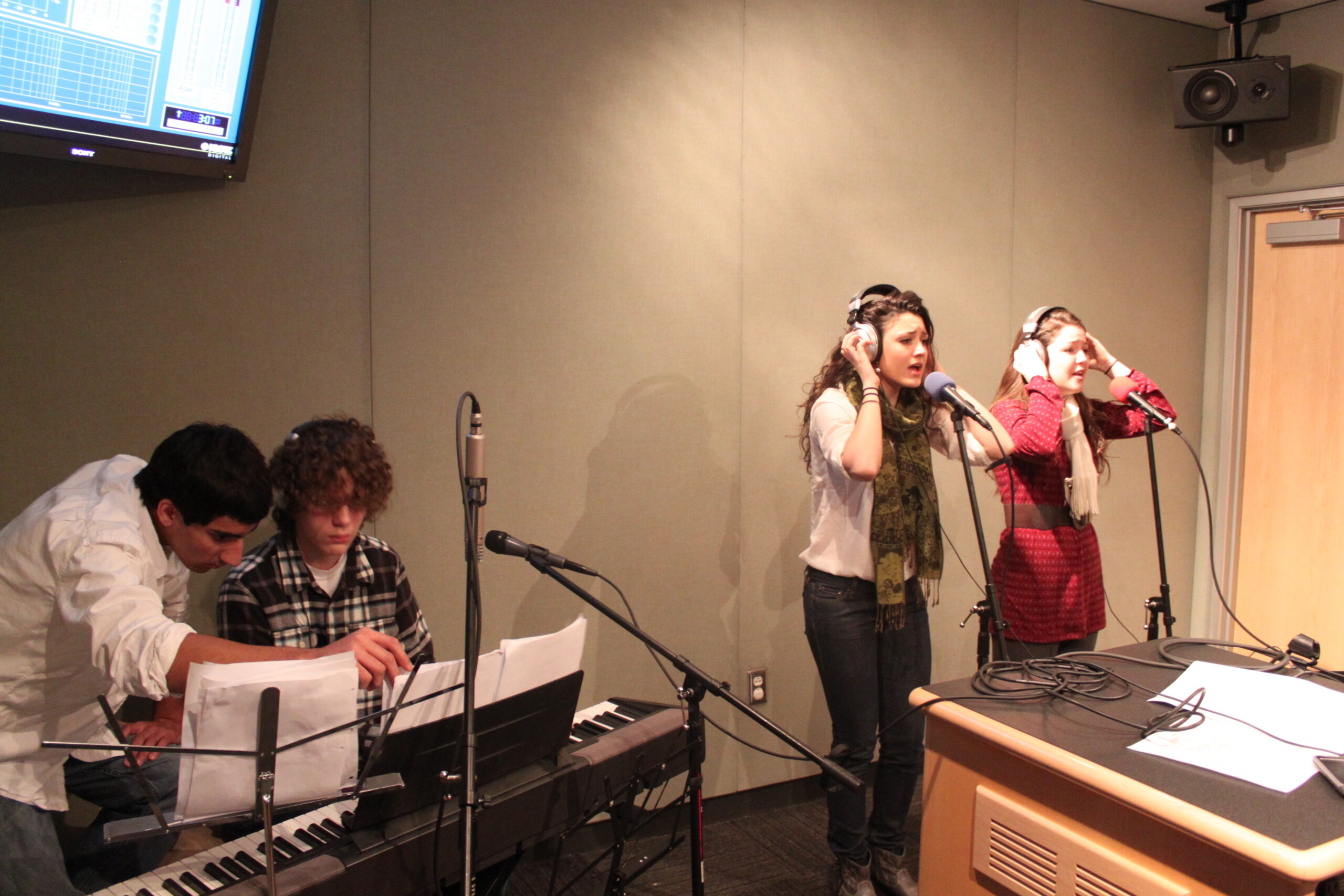 Two teens wearing headphones singing into a microphone and two teens behind a piano keyboard in a radio studio