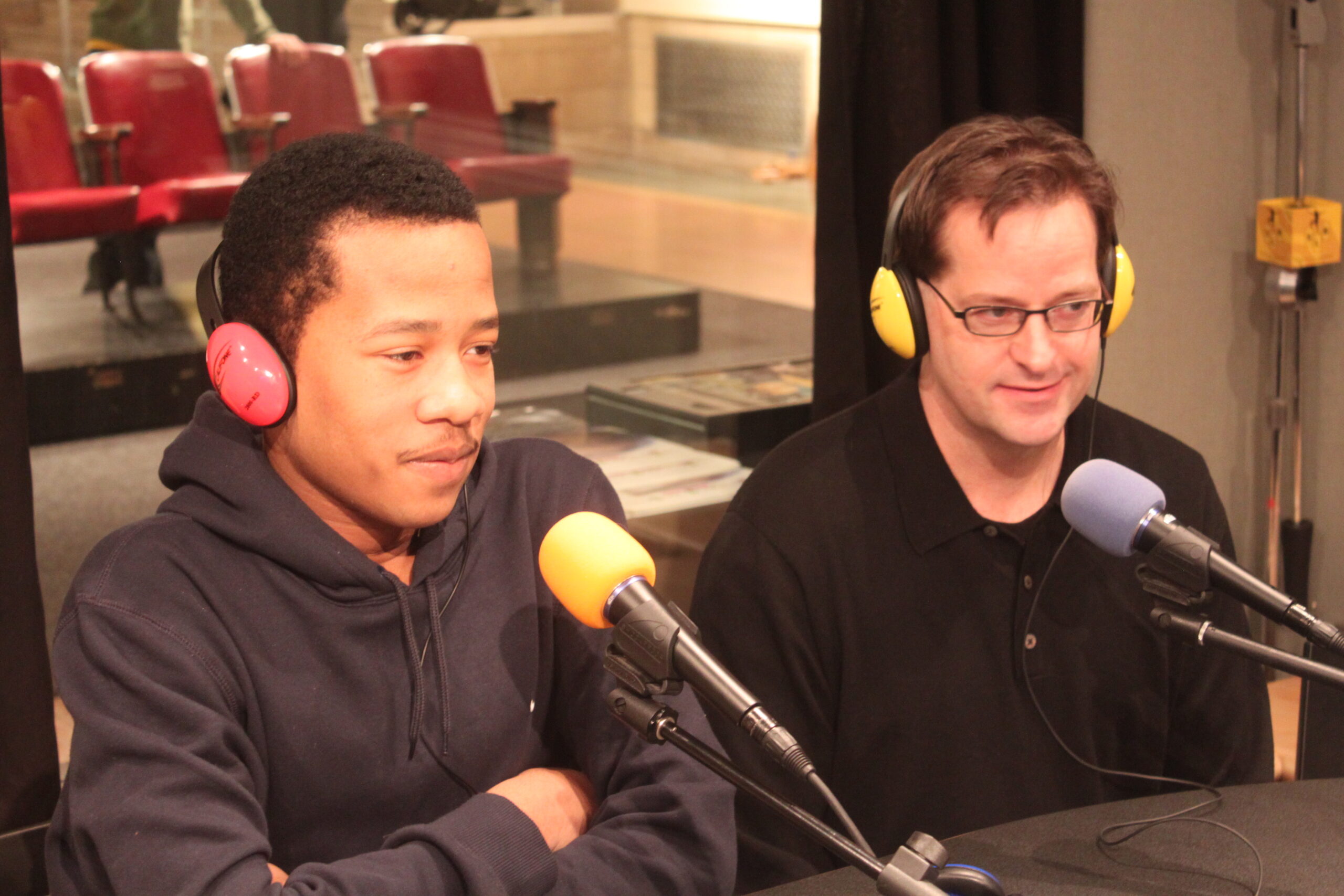 A teen and an adult wearing headphones sitting behind microphones in a radio studio