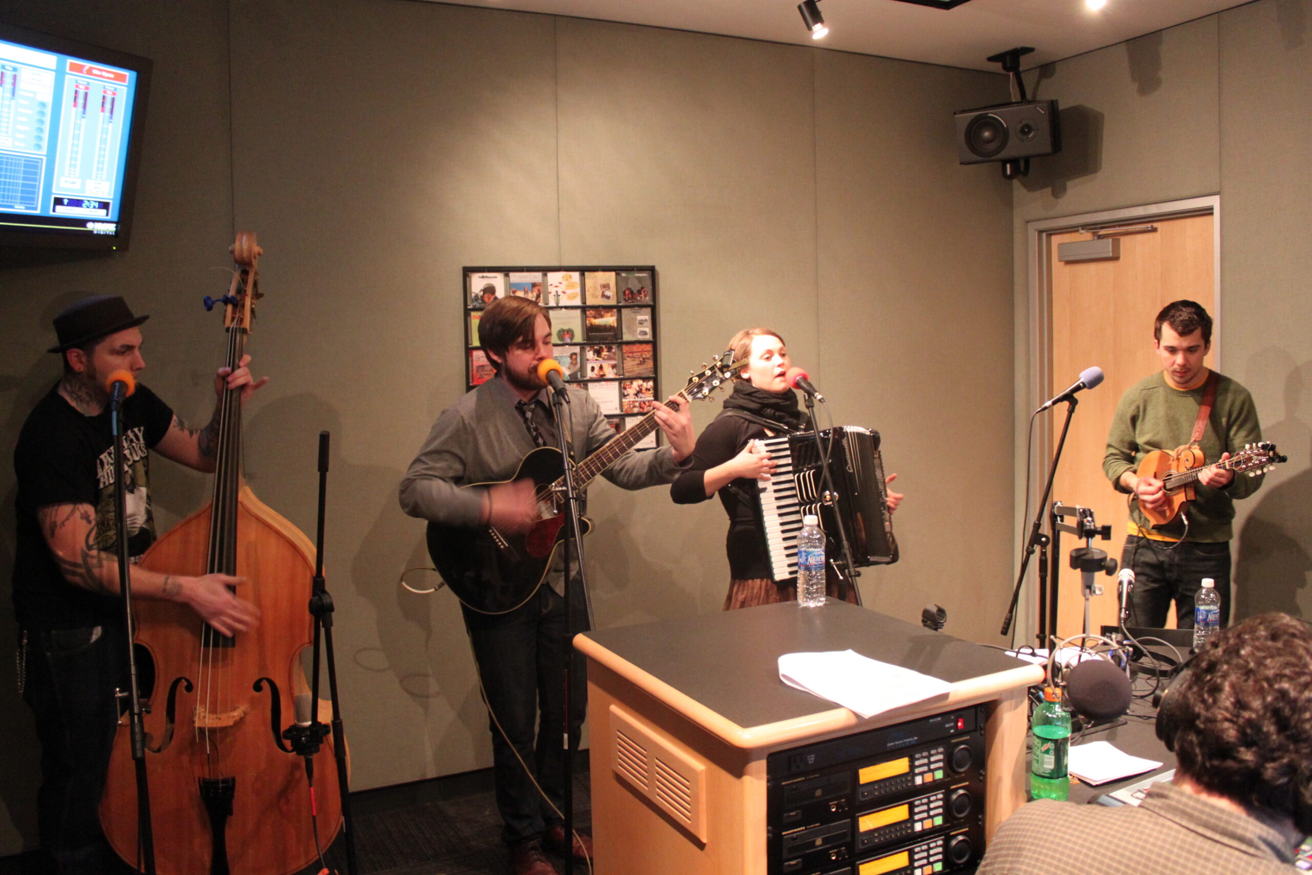 An adult playing upright bass, an adult playing acoustic guitar, an adult playing accordion, and an adult paying ukulele in a radio studio