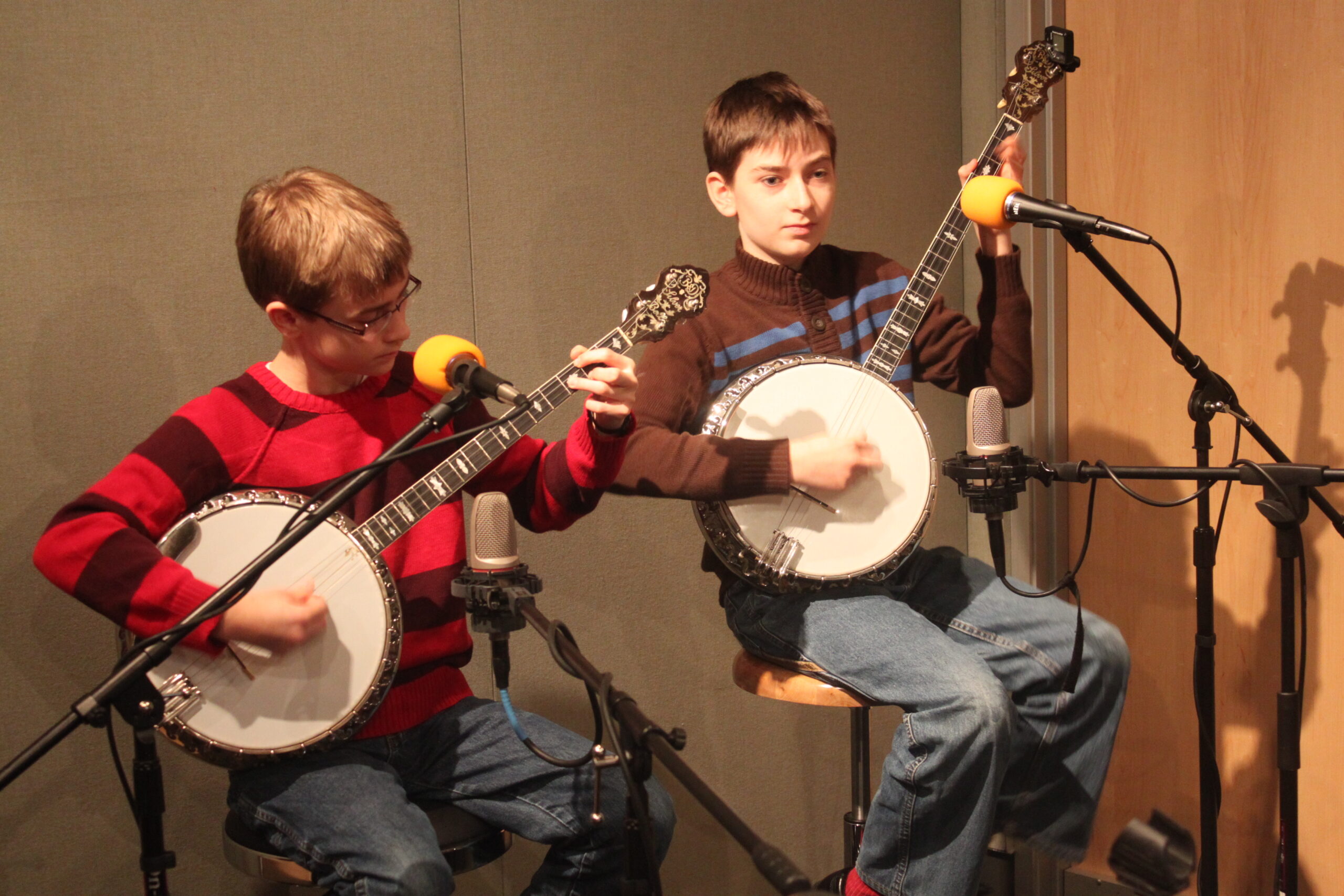 Two youth playing banjos sitting behind microphones in a radio studio