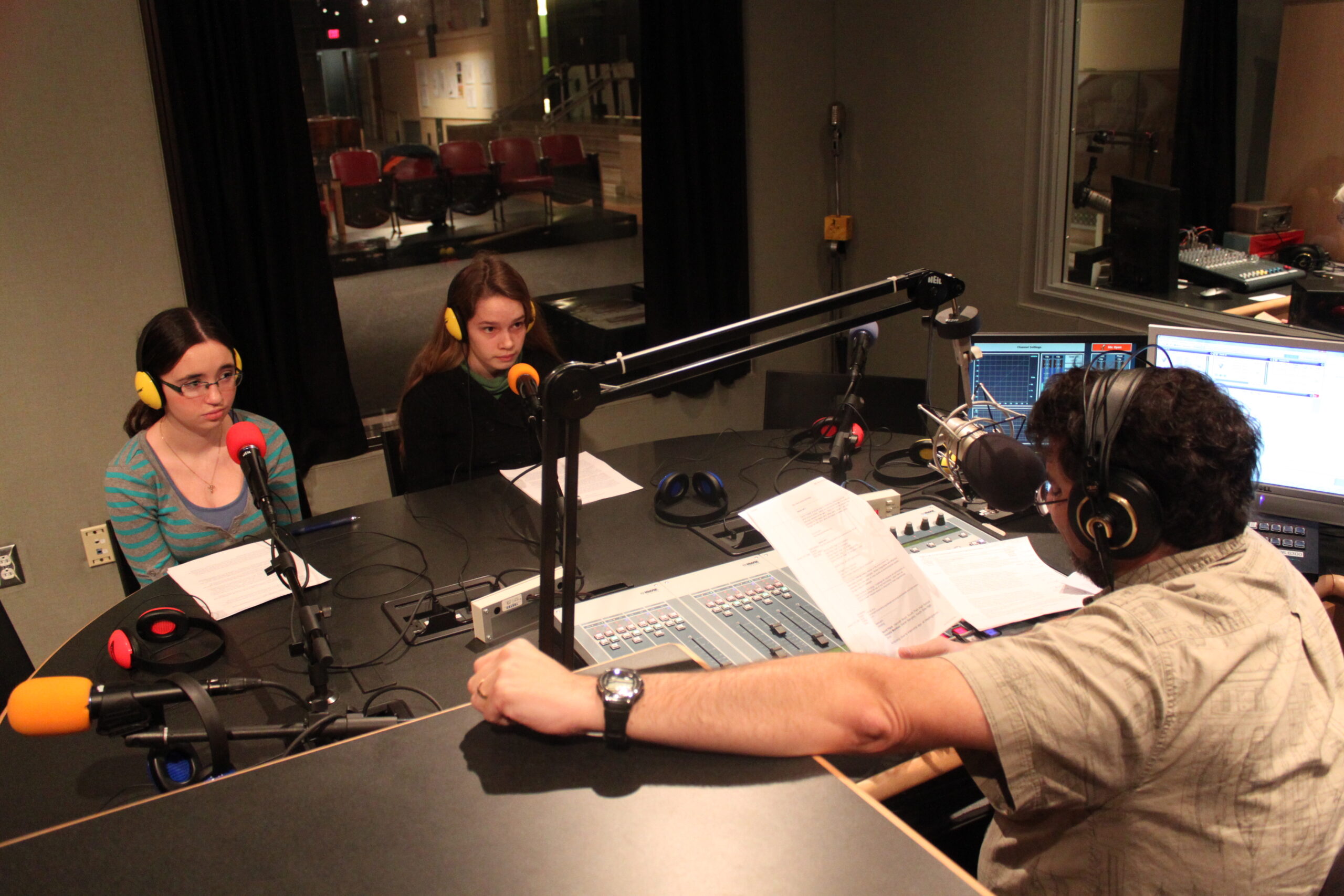 Two teens wearing headphones sitting behind microphones being interviewed by an adult wearing headphones and sitting behind a microphone and an audio mixer in a radio studio