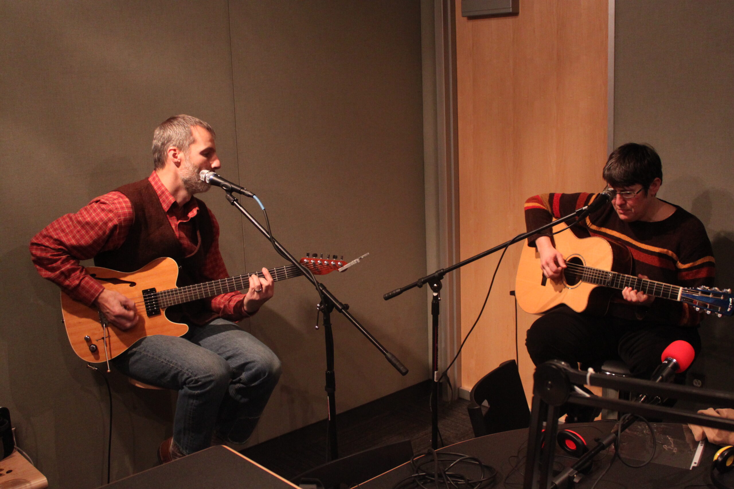 An adult playing acoustic guitar and an adult playing electric guitar, both sitting behind microphones in a radio studio