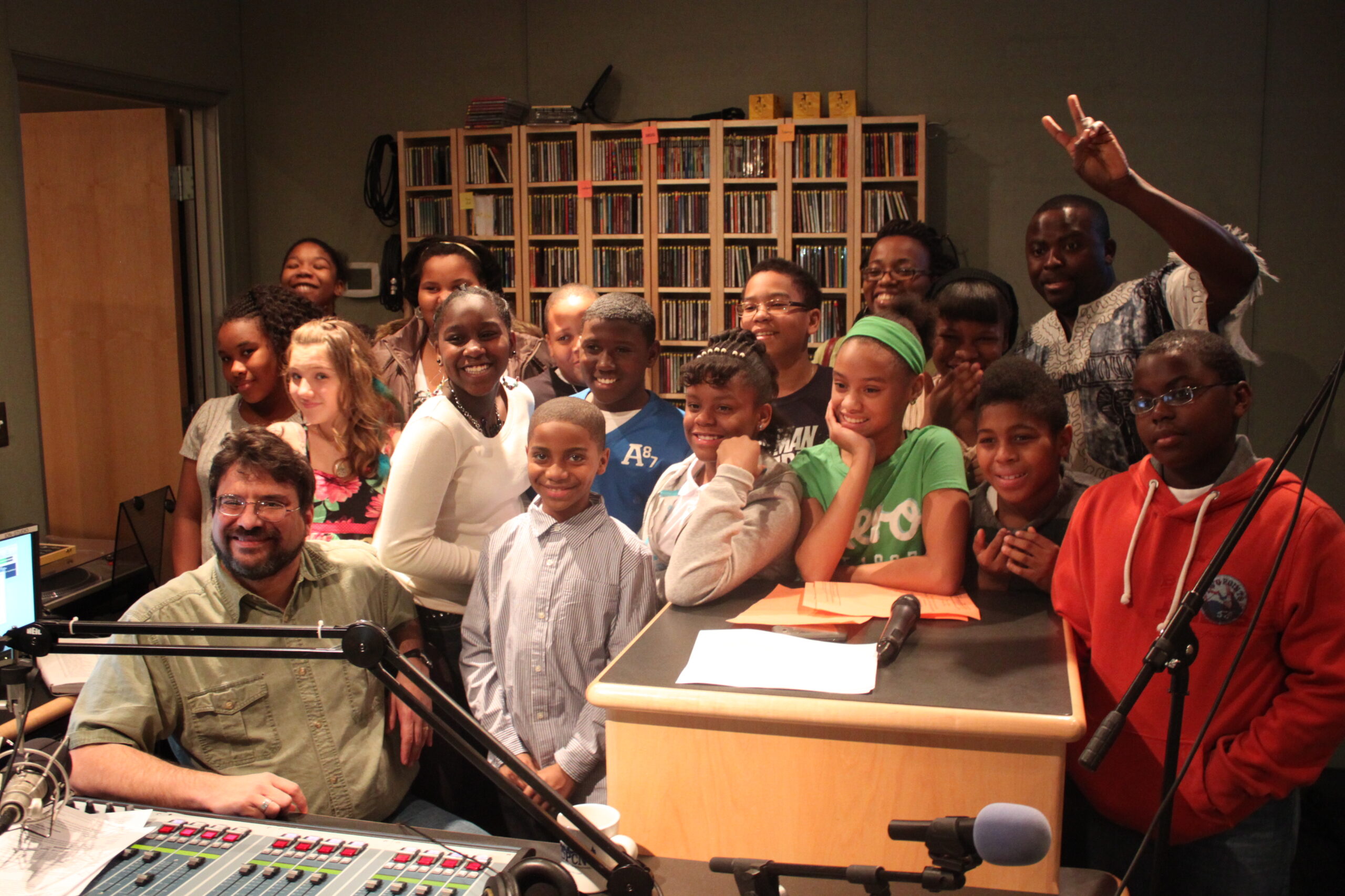 Sixteen youth and an adult smiling and posing in profile in a radio studio