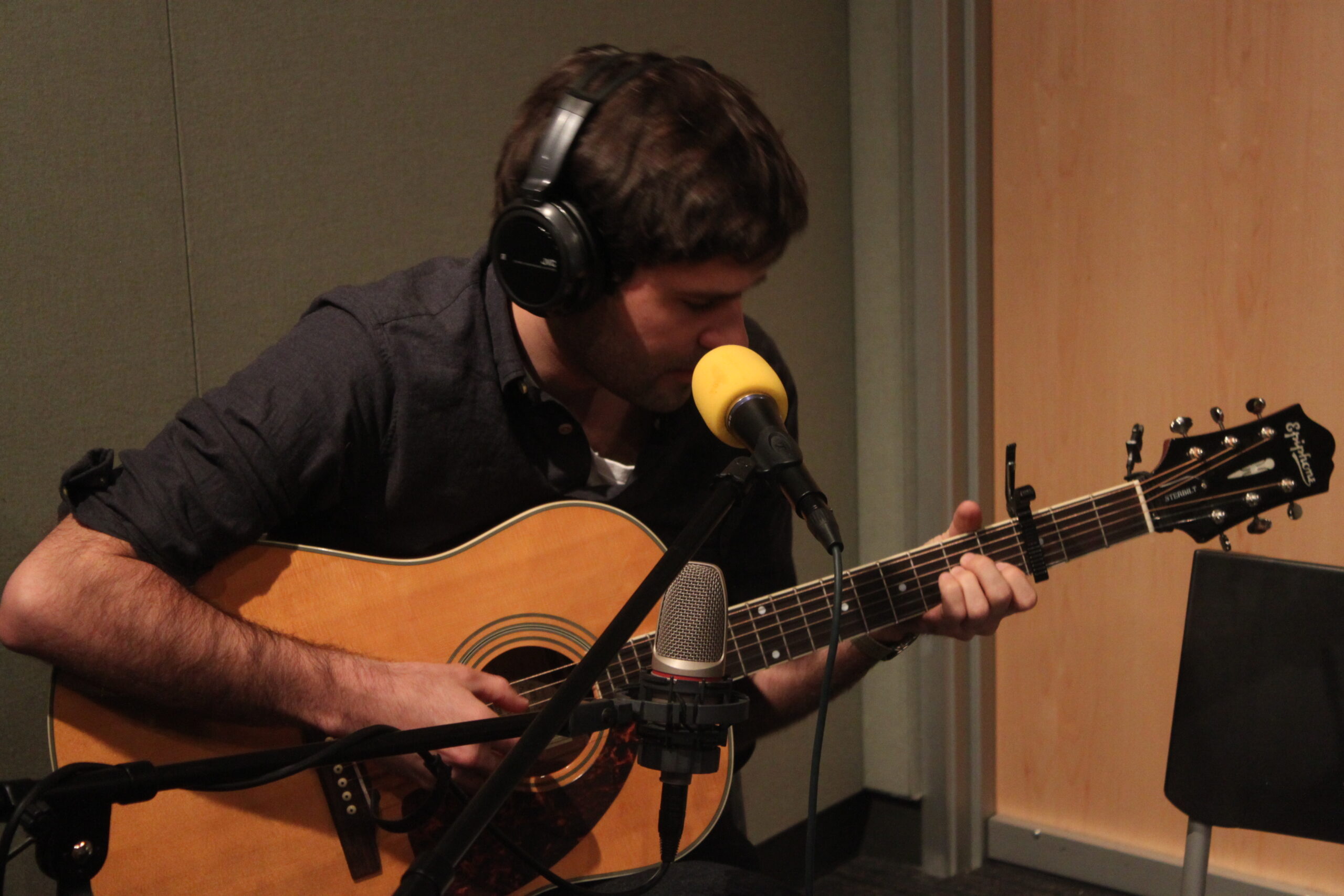 An adult wearing headphones sitting and playing acoustic guitar in a radio studio